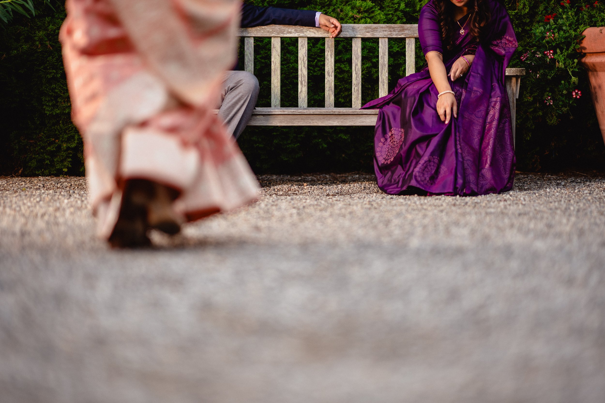 NJ Wedding Photography of A woman in a purple traditional dress sitting on a wooden bench, with a person in a dark suit sitting next to her, and another person walking in the foreground wearing a pink dress,