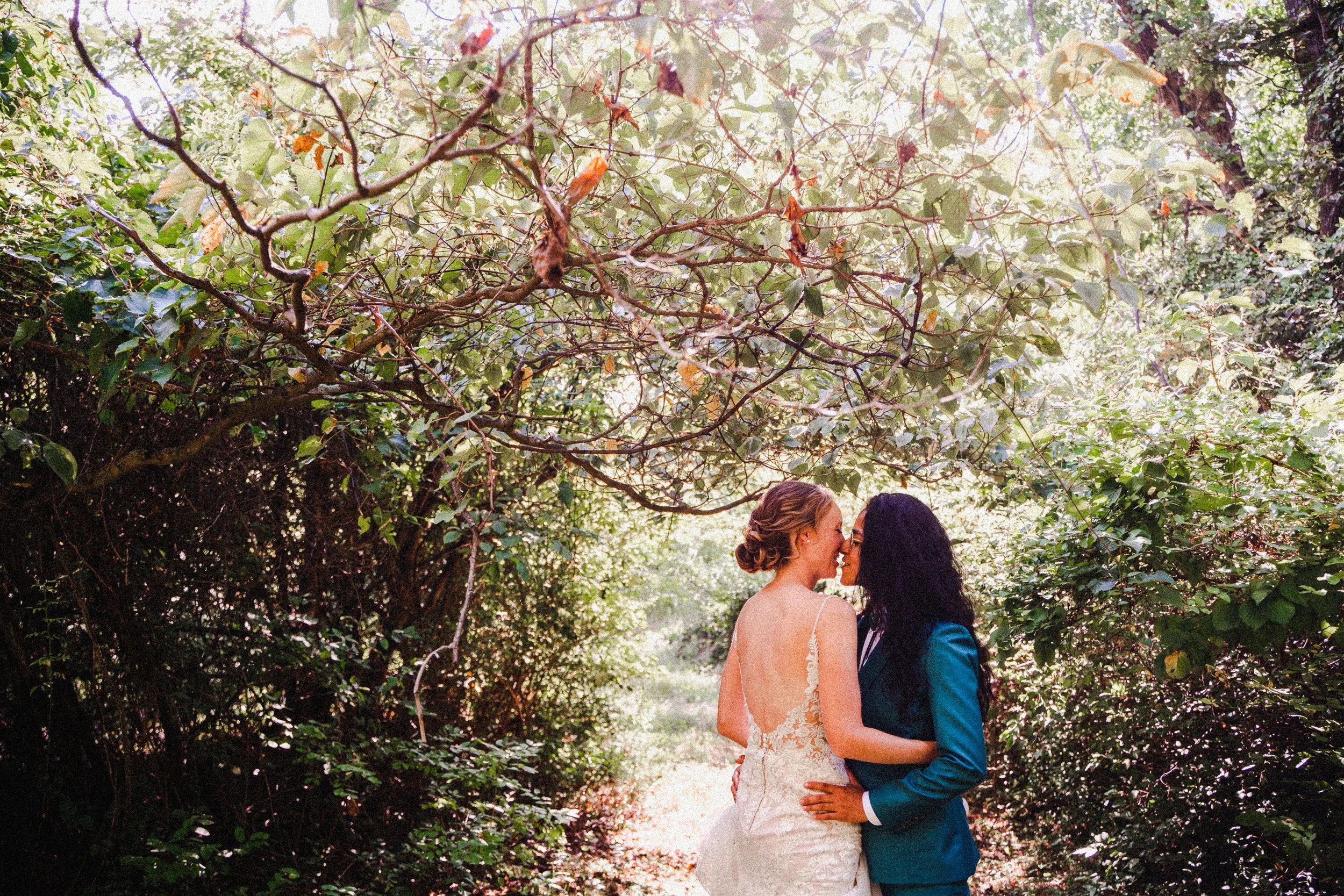 Two women in elegant wedding dresses stand together in a serene forest setting, surrounded by trees and natural beauty.