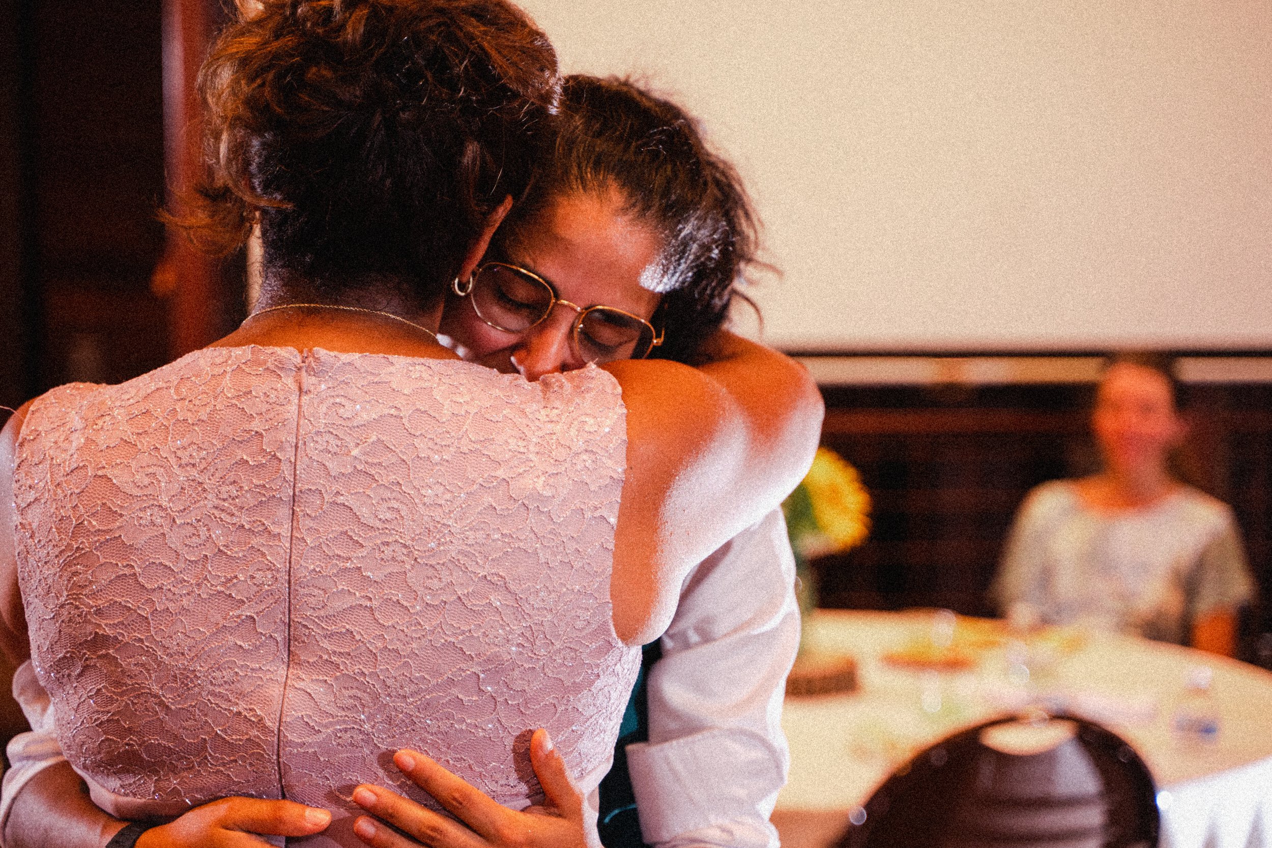A woman embraces another woman joyfully at a wedding celebration, surrounded by festive decorations.