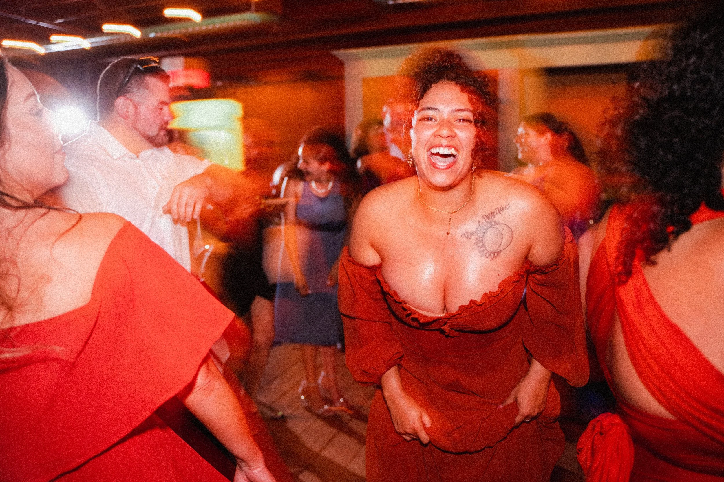 A woman in a red dress laughs joyfully at a lively party, surrounded by festive decorations and smiling guests.