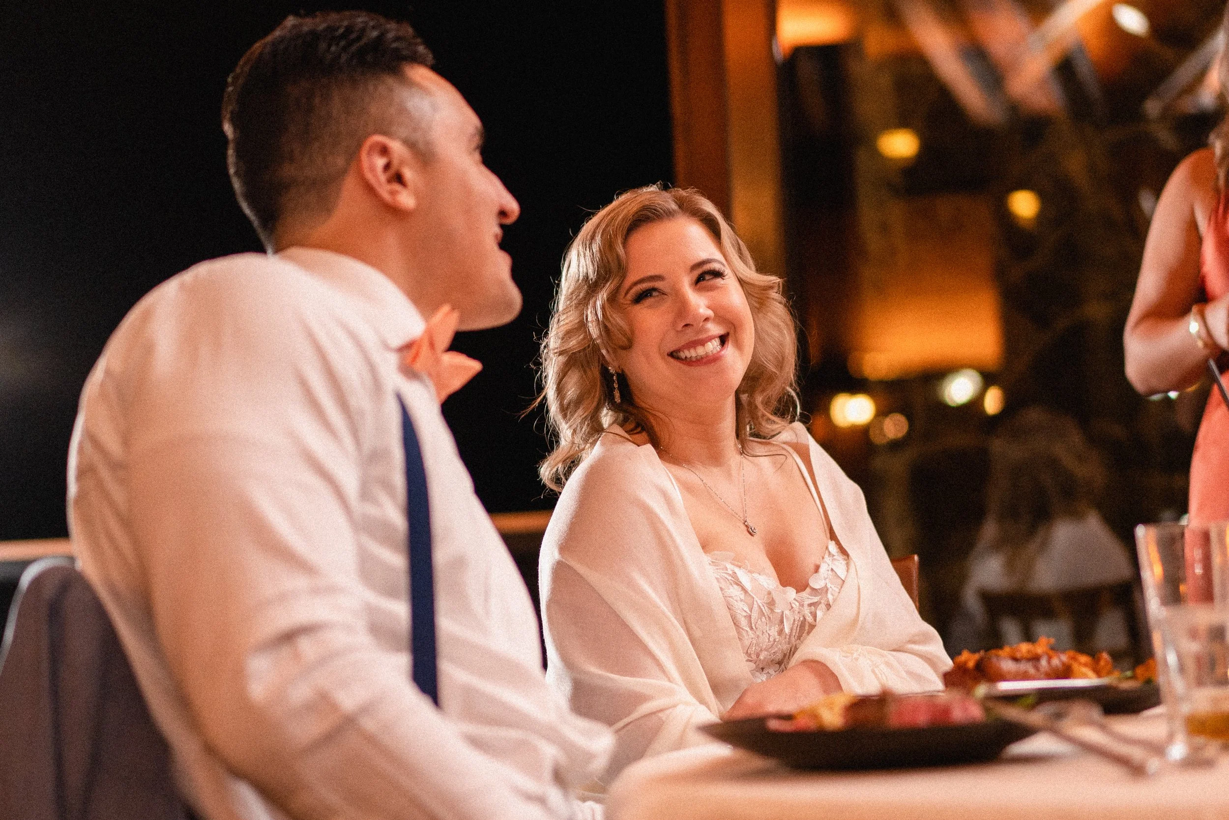 A bride and groom share a joyful smile at each other during their wedding reception, surrounded by festive decorations.