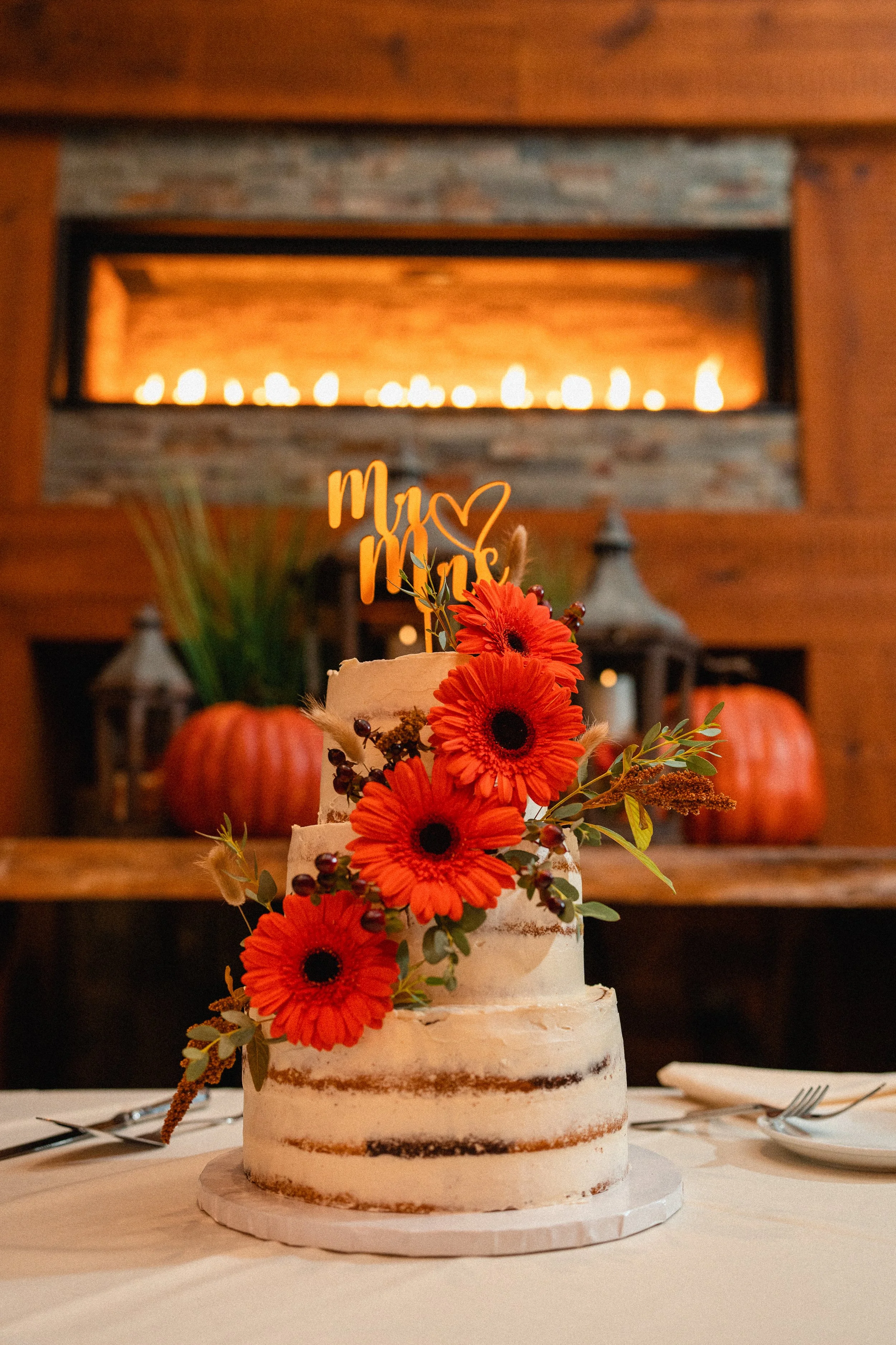 A wedding cake adorned with vibrant orange flowers and a rustic wooden topper, elegantly displayed on a white tablecloth.