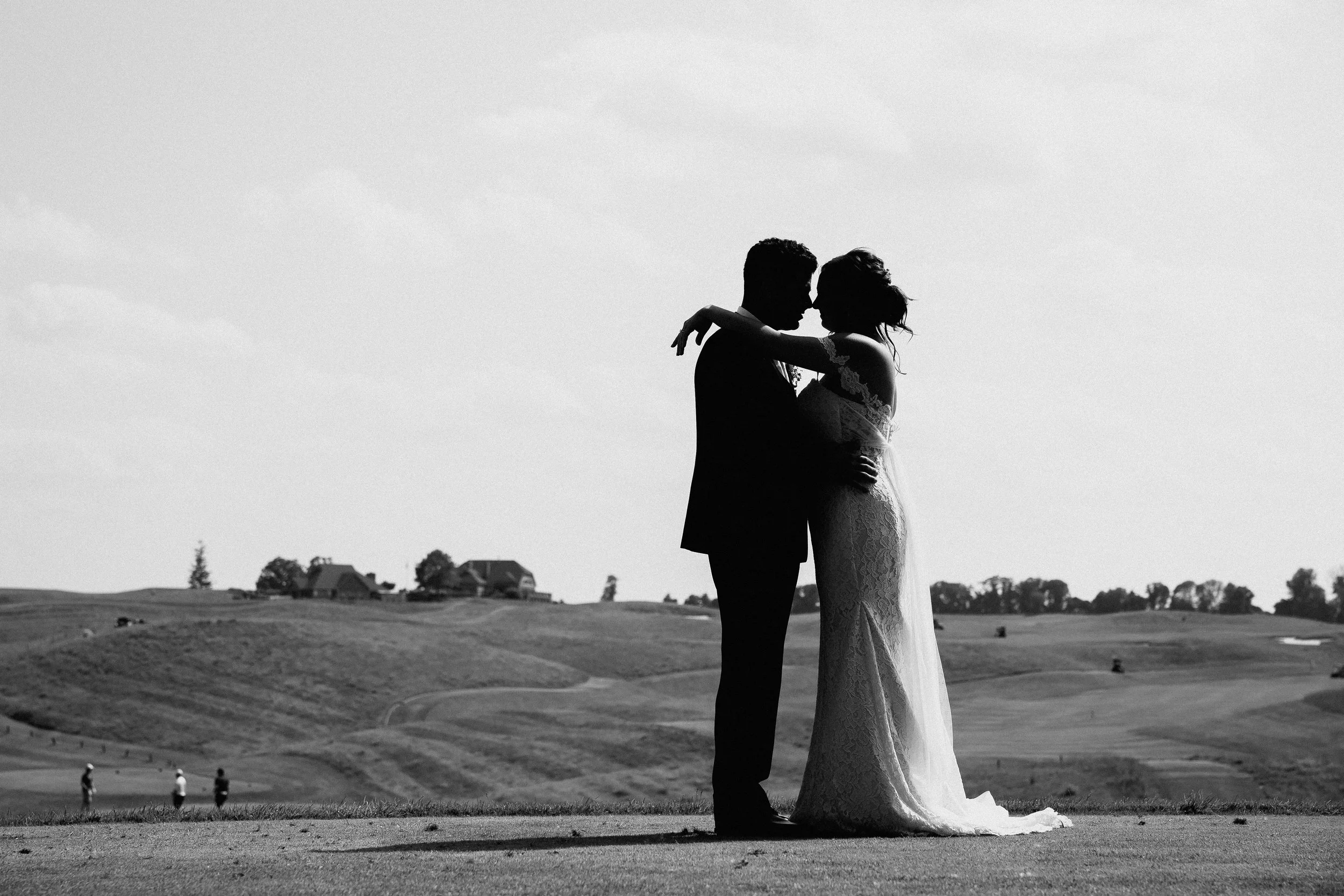 A bride and groom embrace on a lush golf course, surrounded by greenery and a clear blue sky.