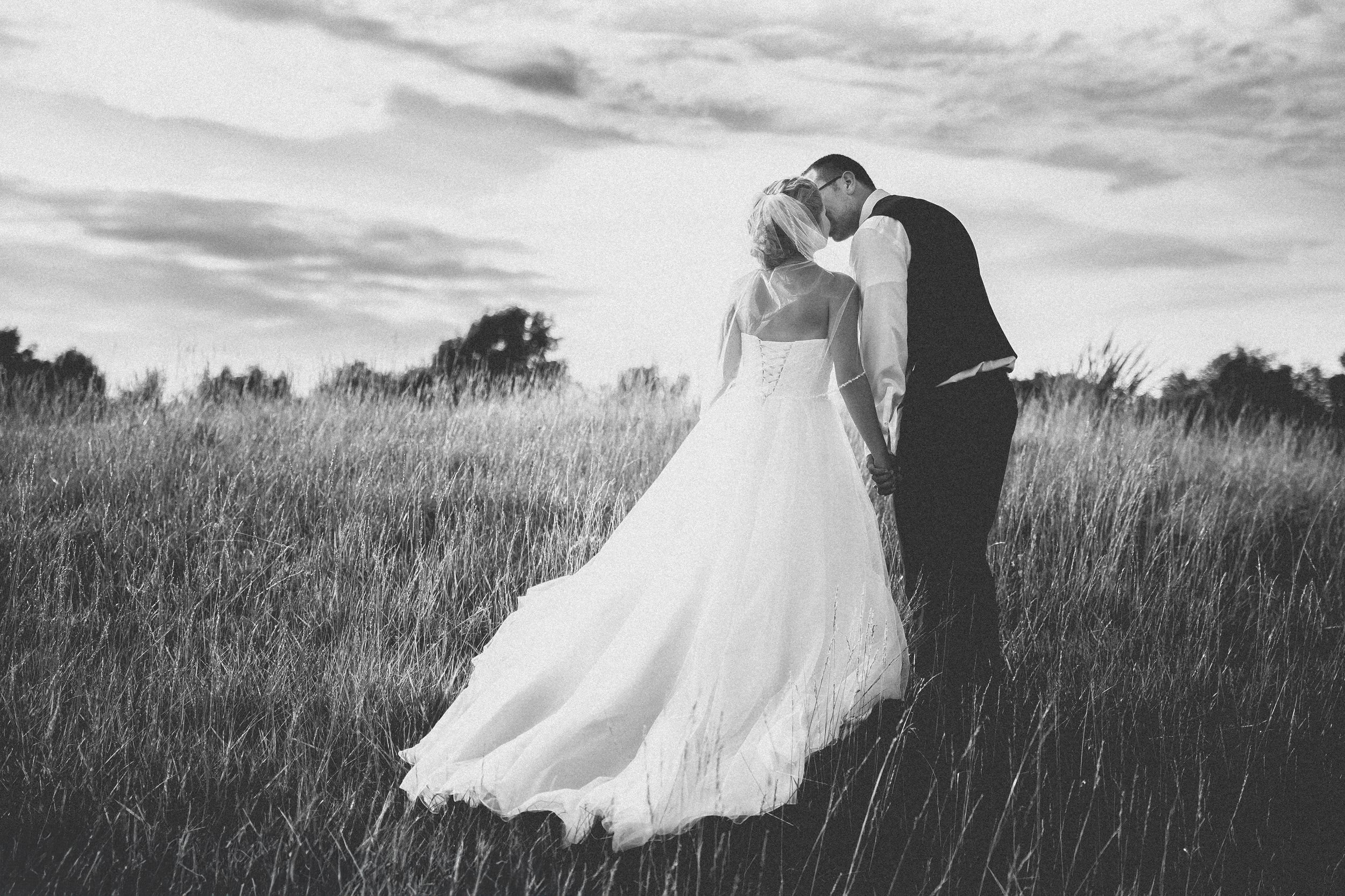 A bride and groom stand together in a field under a cloudy sky, capturing a moment of love and celebration.