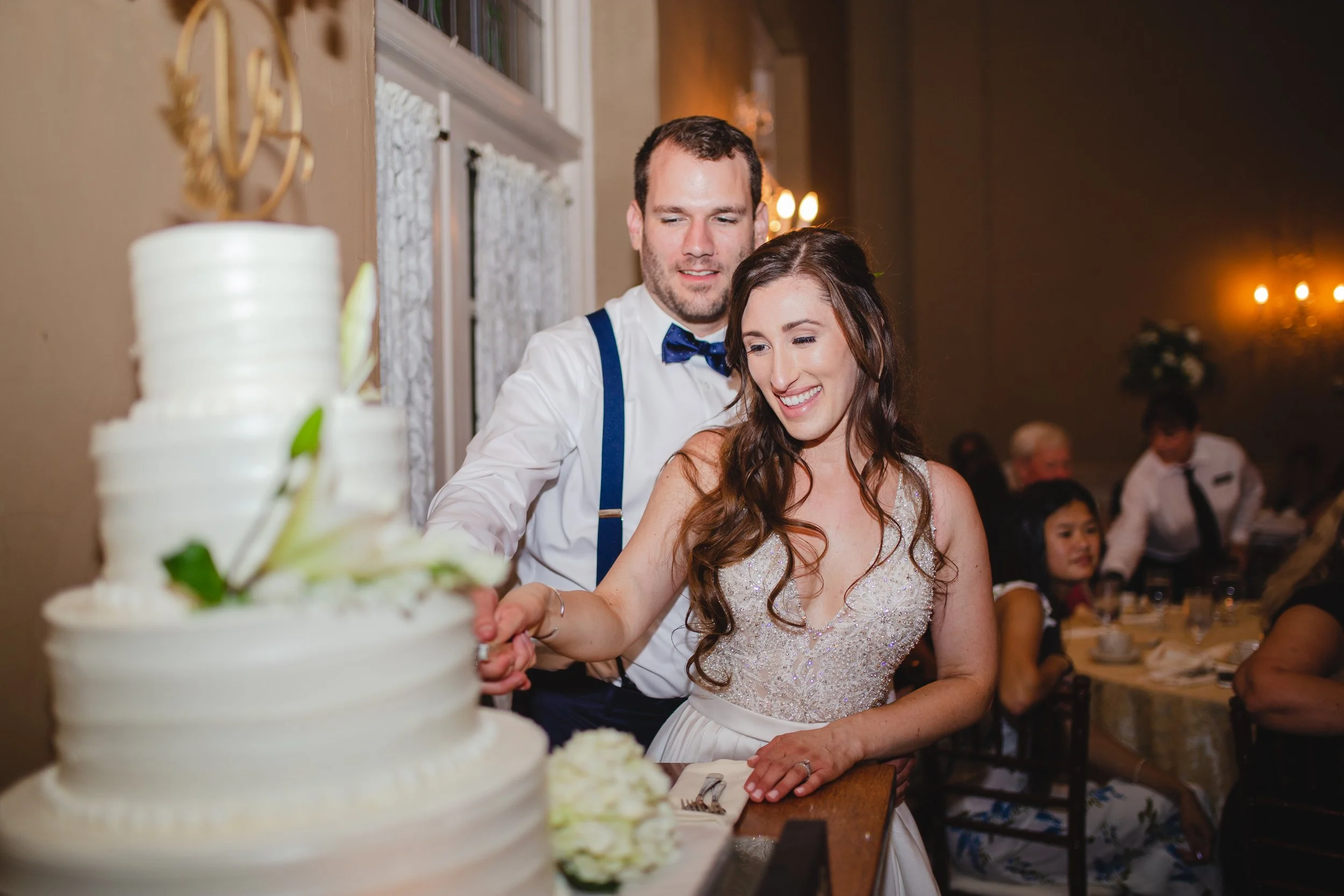 A bride and groom joyfully cutting their wedding cake together, surrounded by floral decorations and smiling guests.