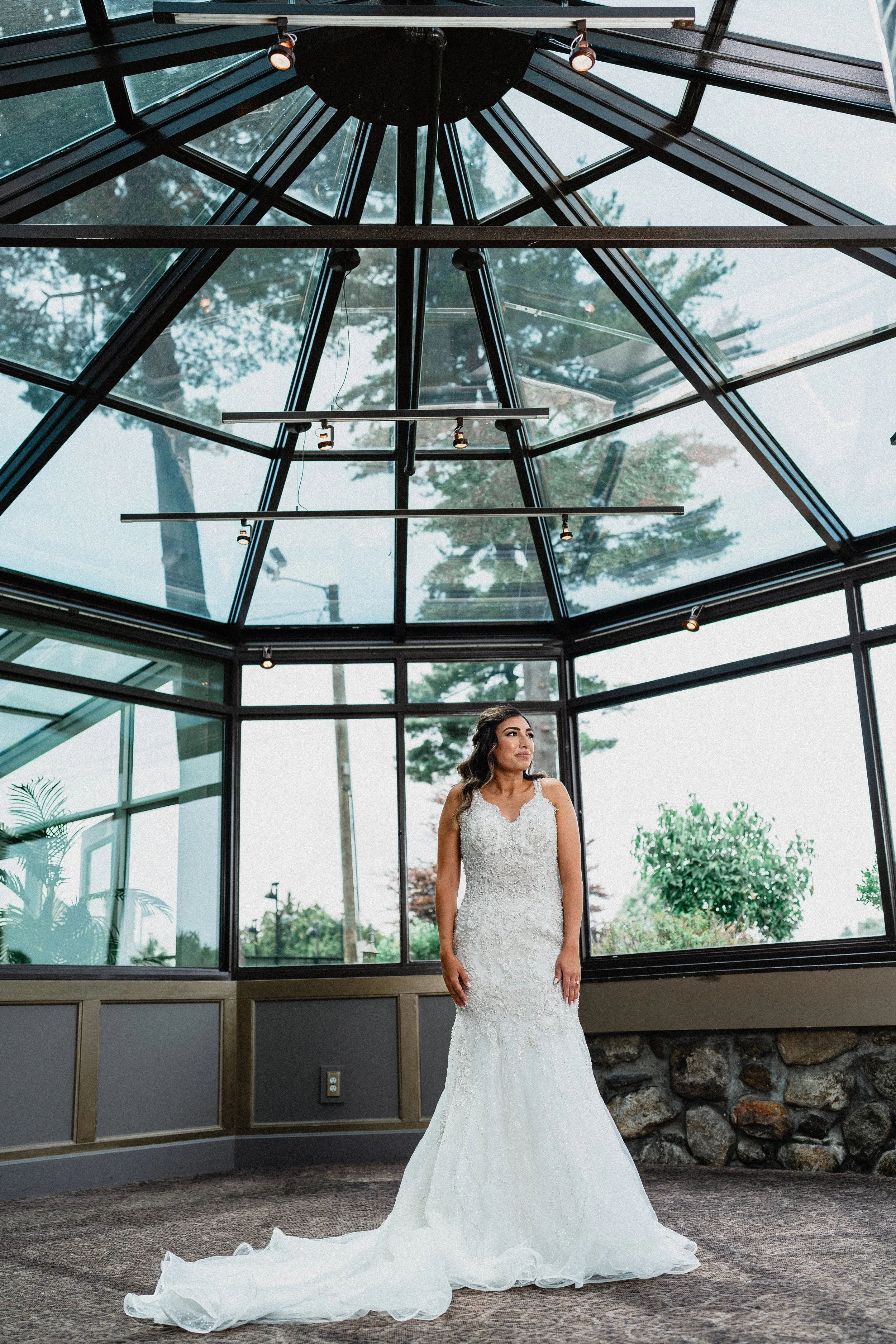 A bride in a white wedding dress stands gracefully in front of a large window, light illuminating her figure.