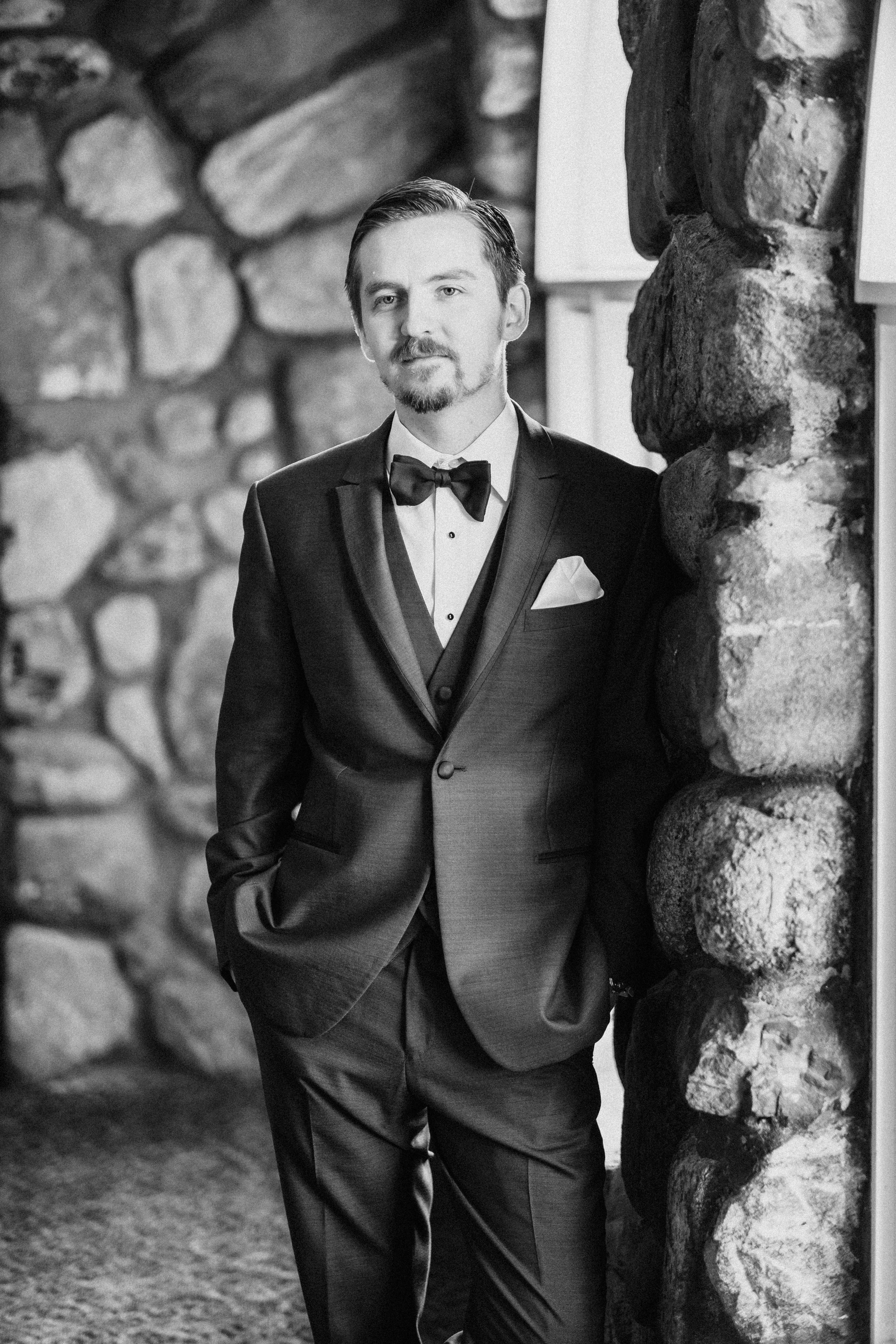 A man in a suit and bow tie casually leans against a wall, exuding confidence and style.