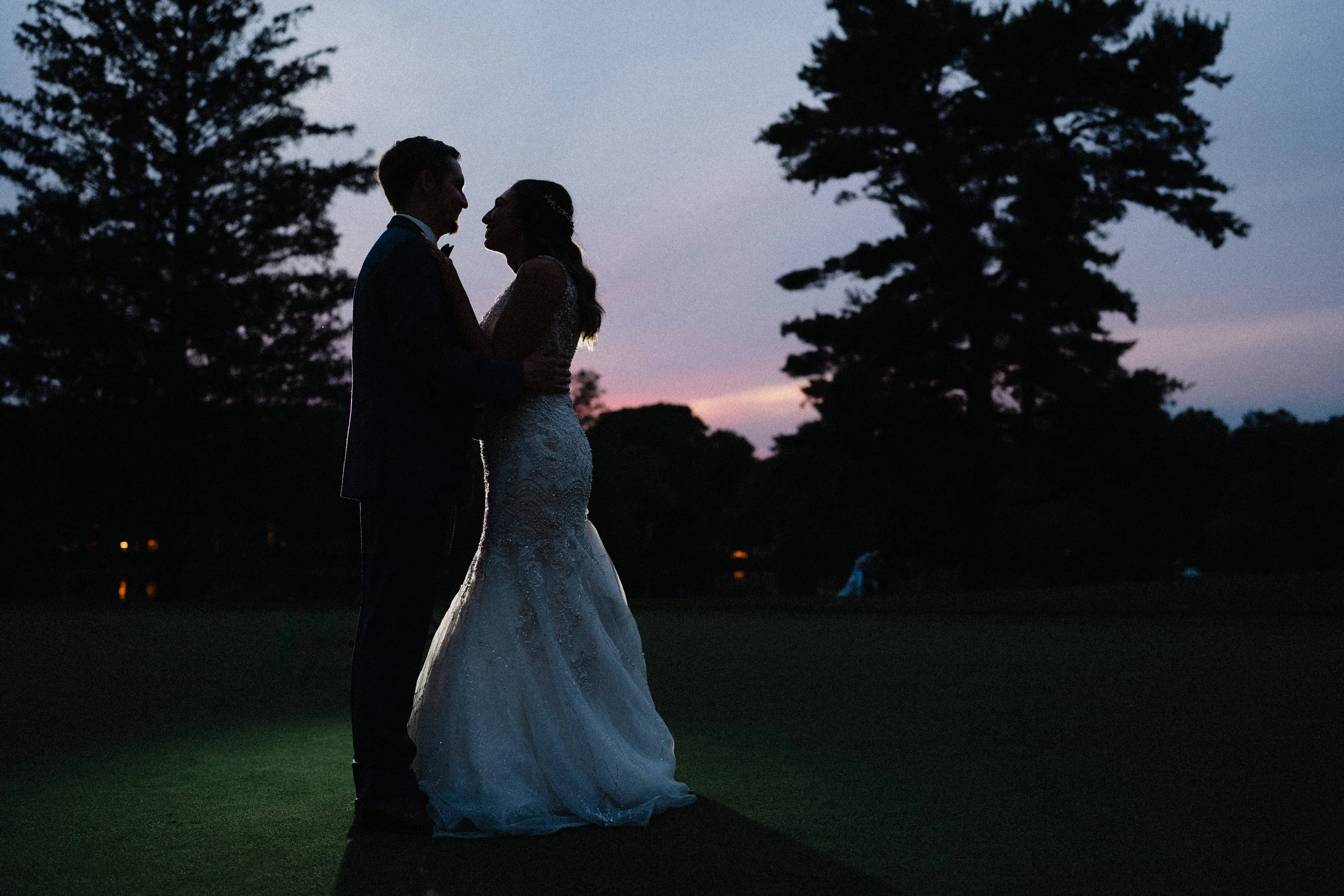 A bride and groom stand together in a grassy field, illuminated by the warm glow of a sunset.