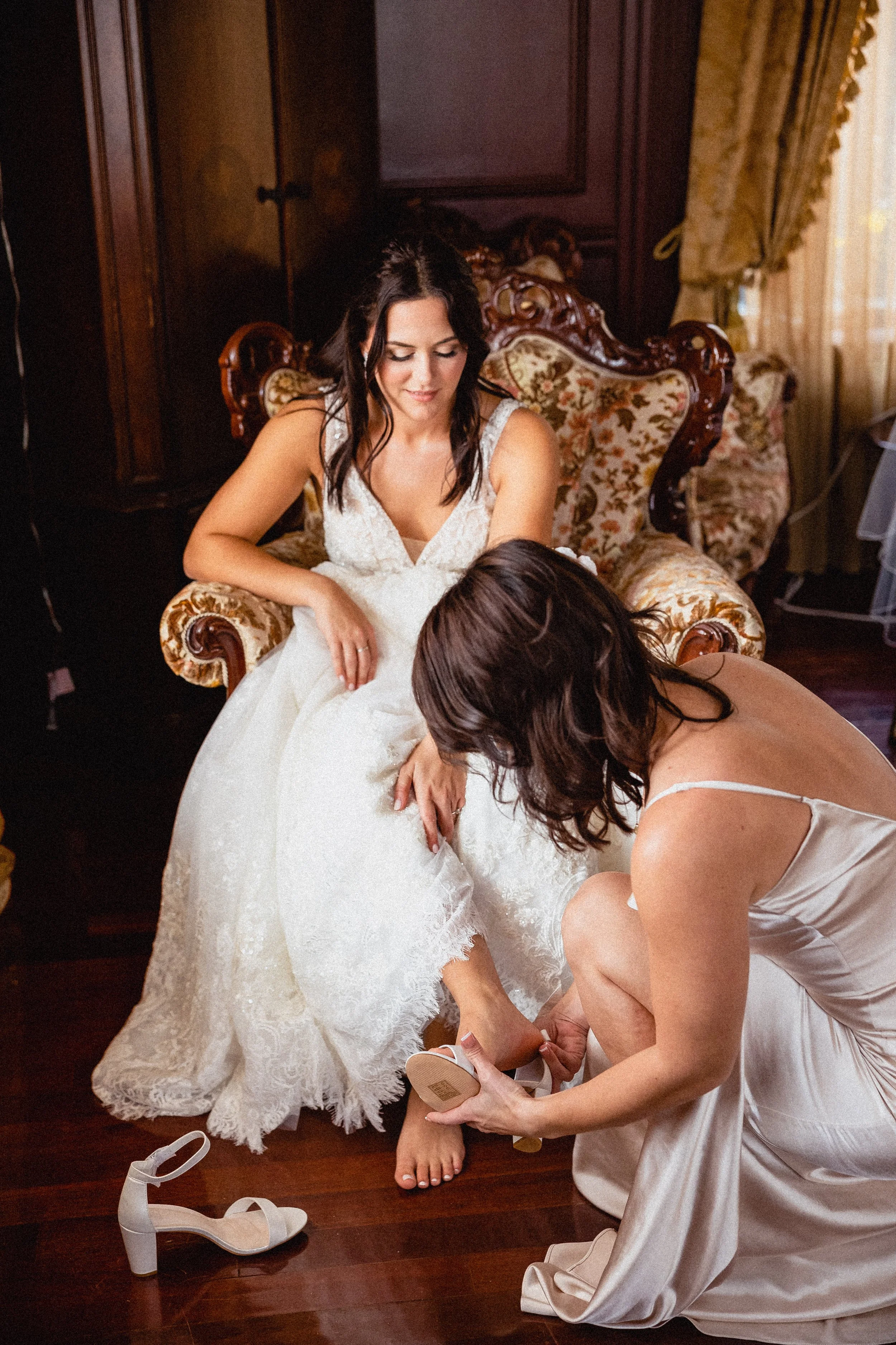 Bride in a white robe, applying makeup in a sunlit room, surrounded by wedding attire and floral decorations.