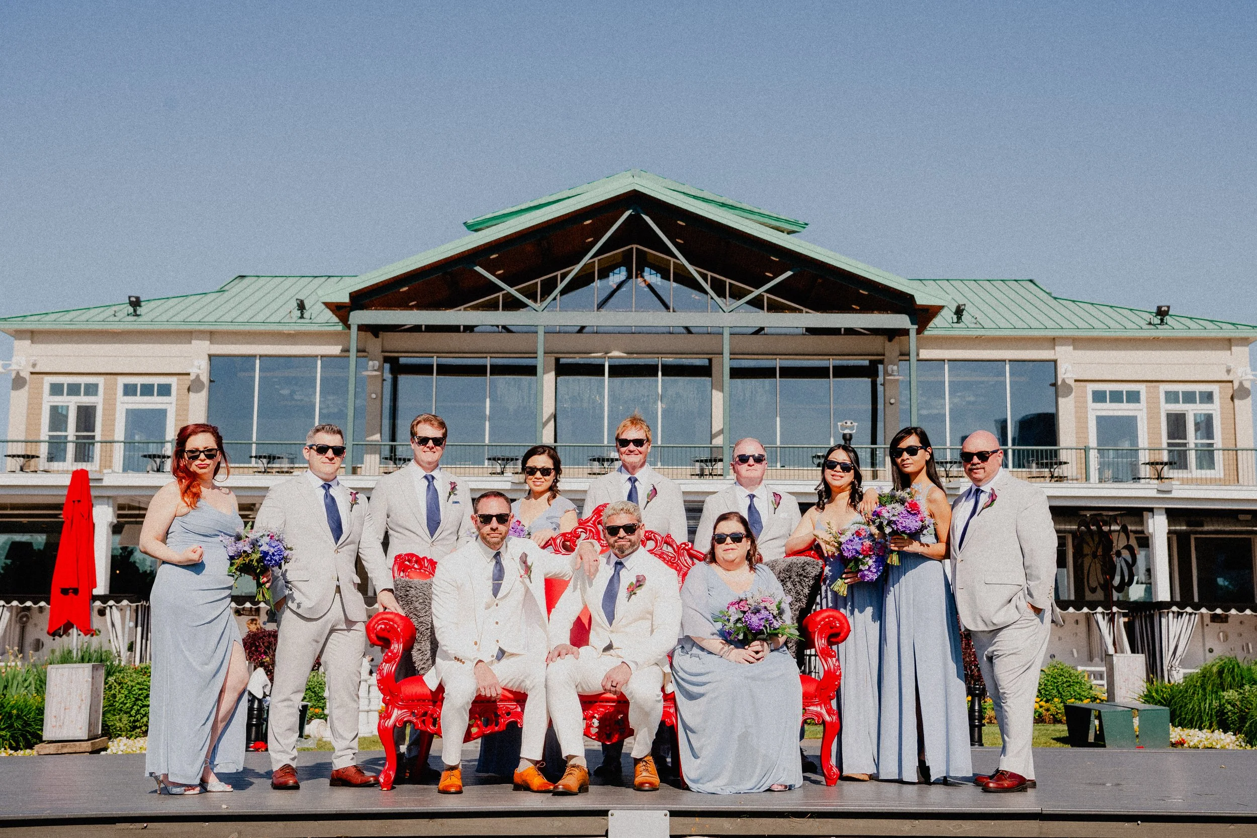 A wedding party smiles for a photo in front of a grand building, dressed in formal attire with flowers in hand.