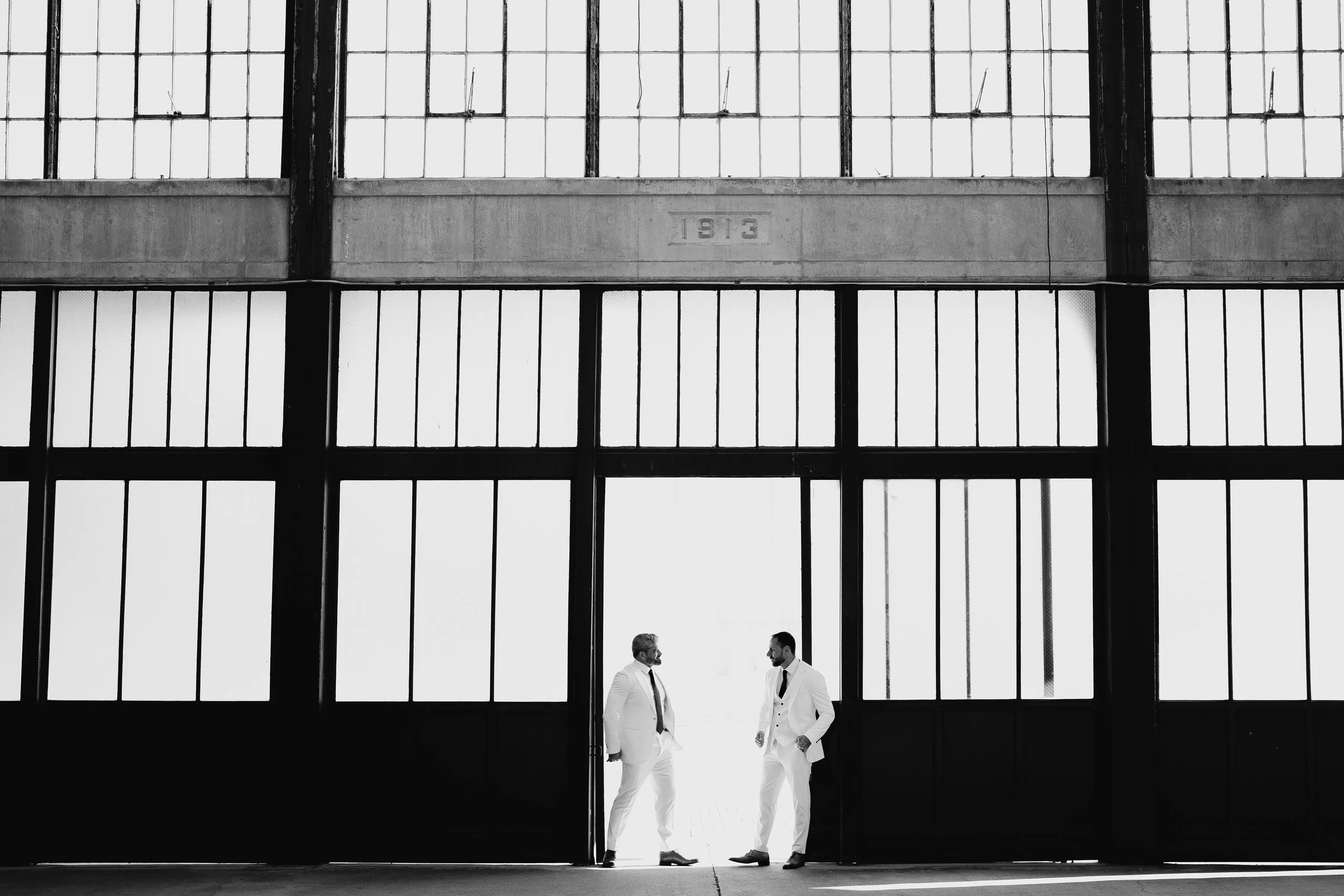 Two men in white suits stand confidently in front of a large, modern building.