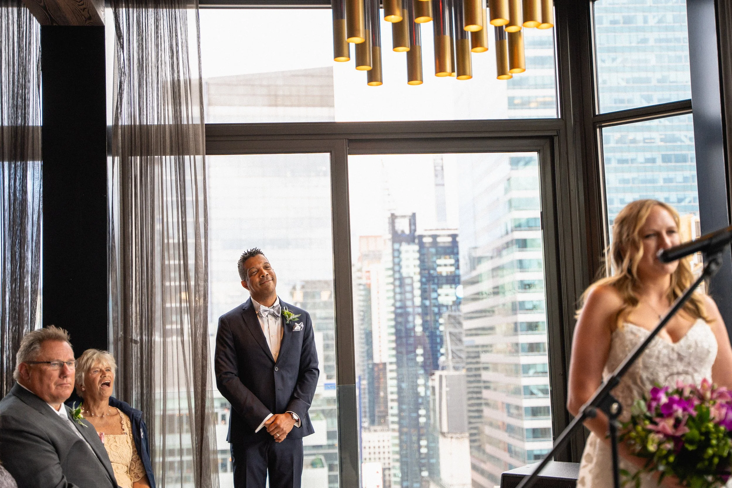 A bride and groom stand together in front of a large window, radiating joy on their wedding day.