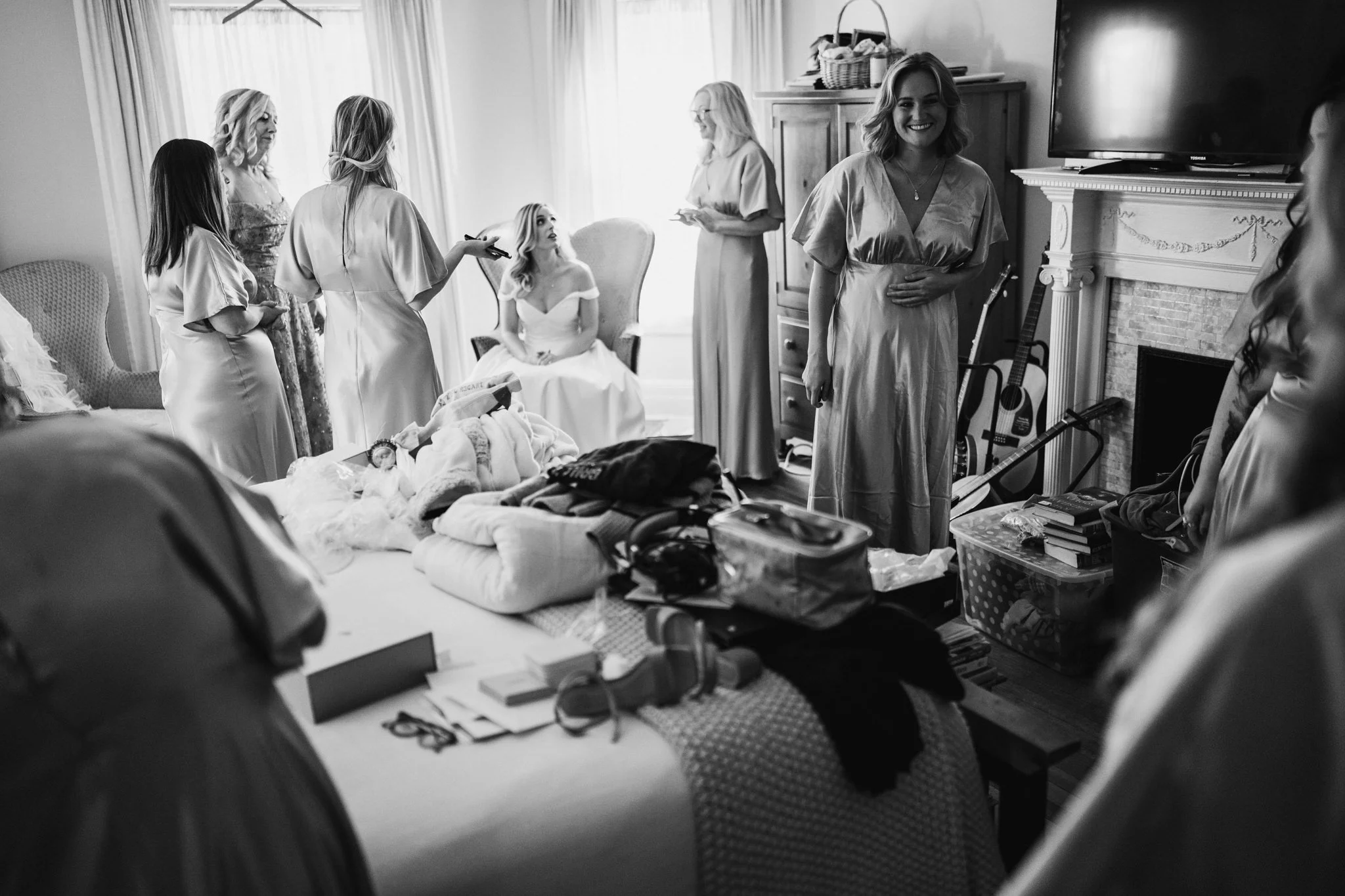 A group of women, likely a bride and her bridesmaids, preparing in a living room with clothes, books, and personal items on a bed. One woman is seated in the background, smiling, while others are standing and talking or using their phones.