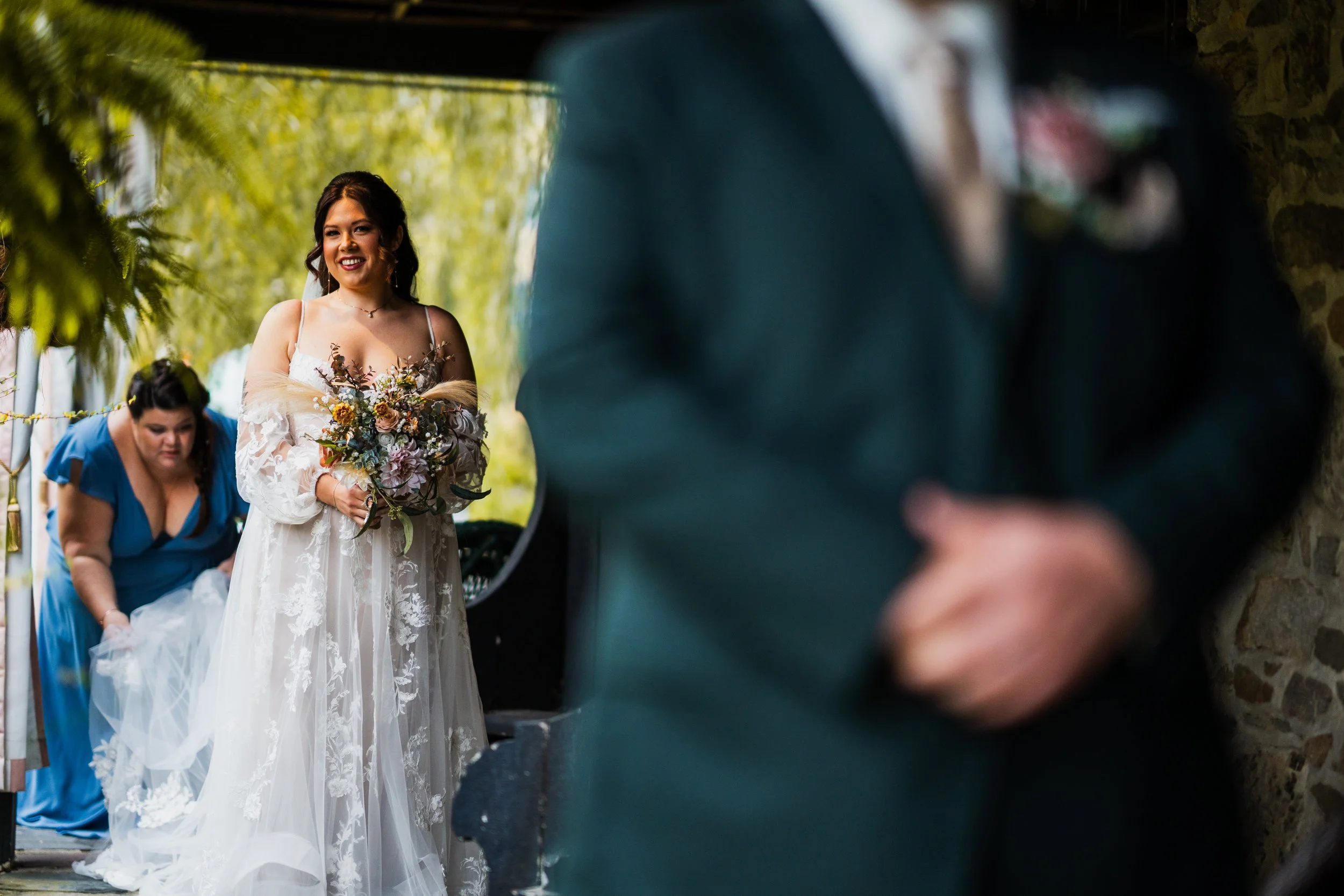 A bride holding a bouquet of flowers, smiling, with a woman in a blue dress adjusting her wedding dress in the background, seen through a blurred reflection of an older man in a dark suit.