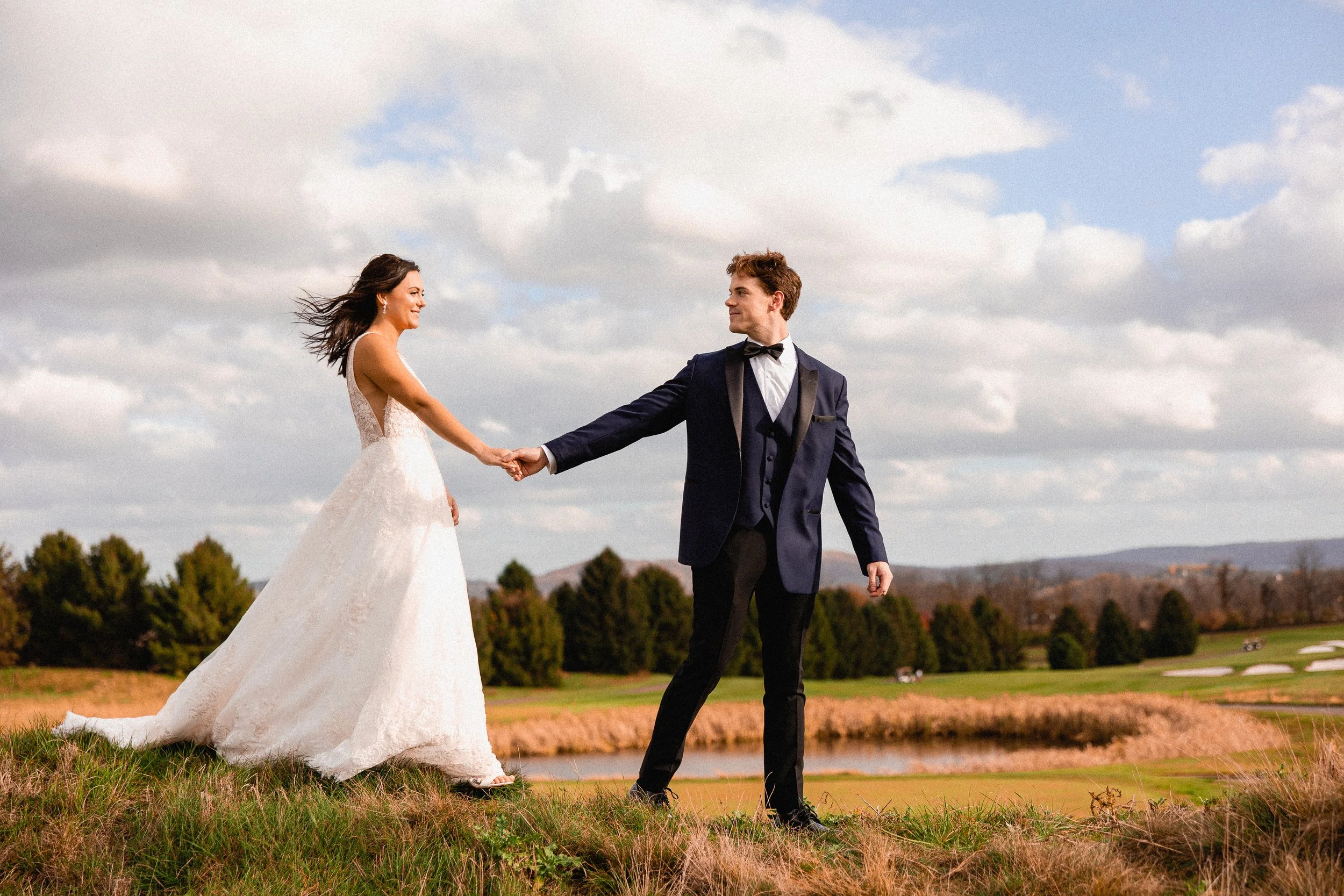 A bride and groom holding hands outdoors on a grassy hill with a golf course and trees in the background, under a partly cloudy sky.