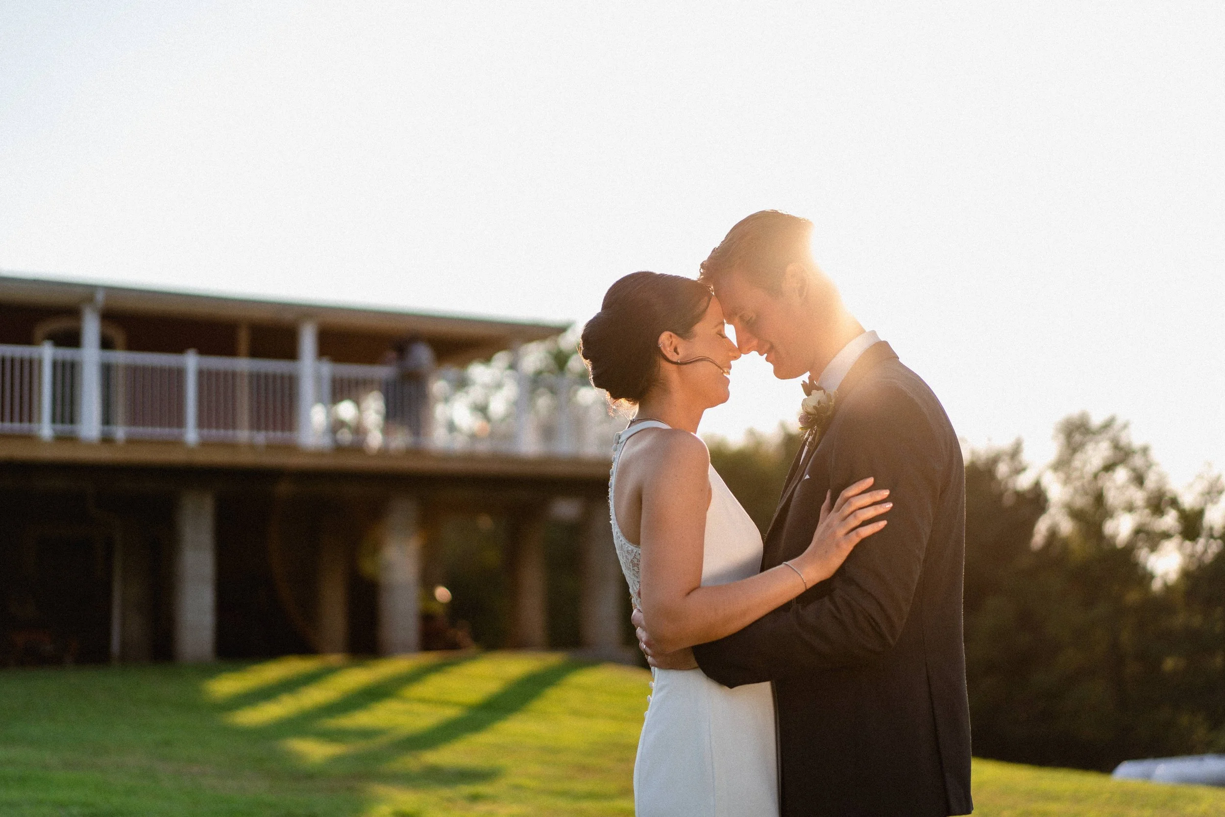 A bride and groom embrace each other with foreheads touching during sunset on a grassy field, with a building in the background.