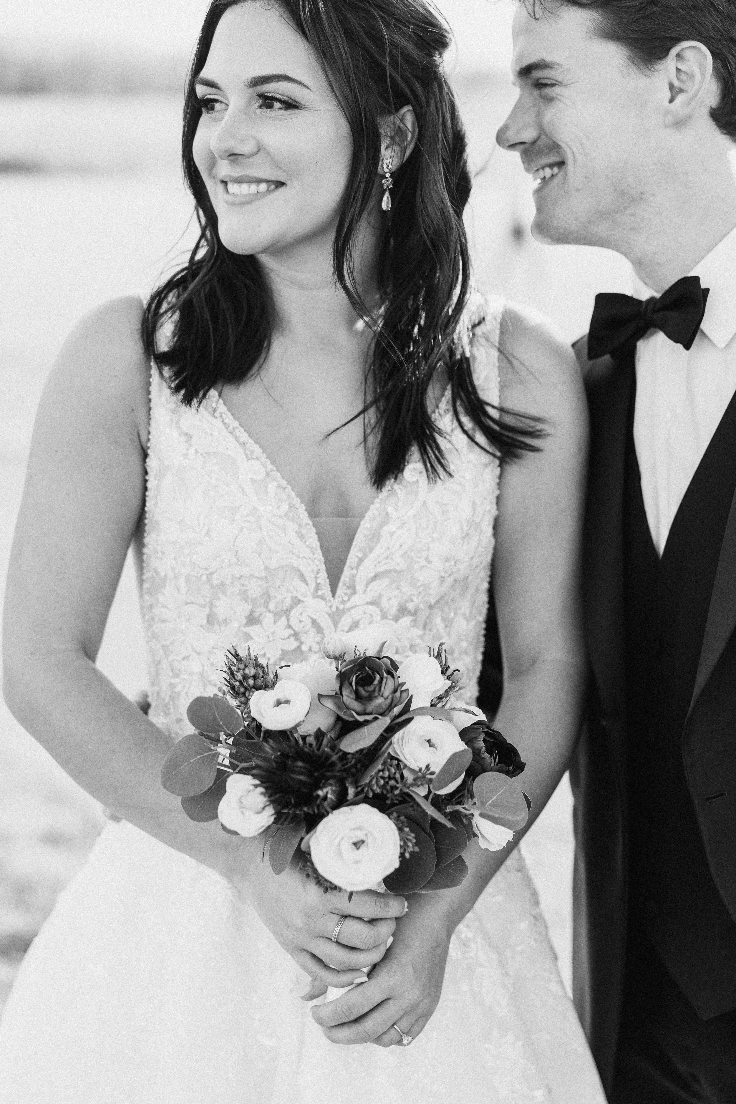 Black and white photo of a bride and groom on their wedding day, smiling at each other. The bride is holding a bouquet of flowers, wearing a lace wedding dress, and the groom is dressed in a tuxedo with a bow tie.