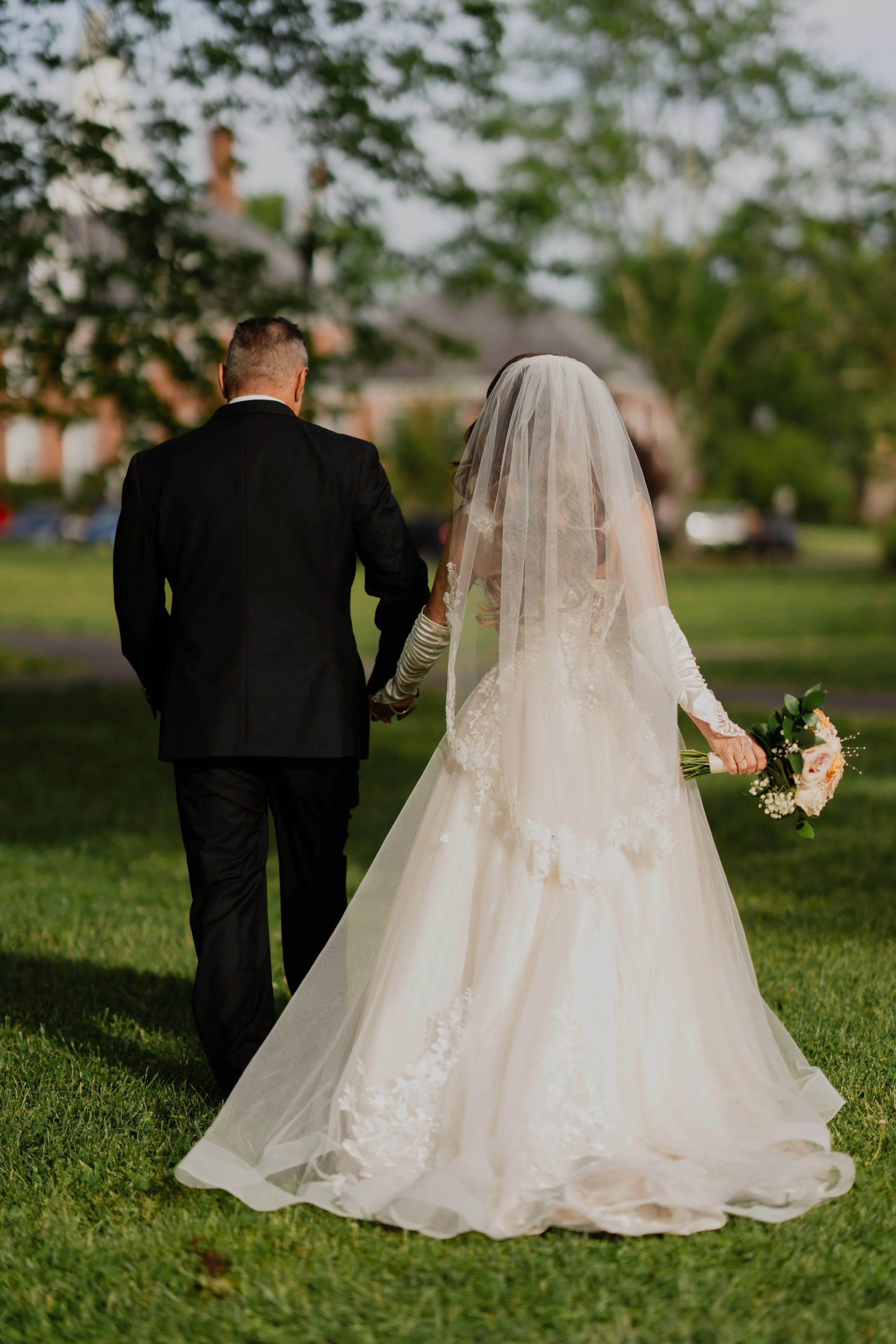 Bride and groom walking outside on a grassy area, holding hands, with the bride holding a bouquet of flowers. The bride is wearing a long white wedding dress with a veil, and the groom is in a black suit. Trees and buildings are visible in the backgr