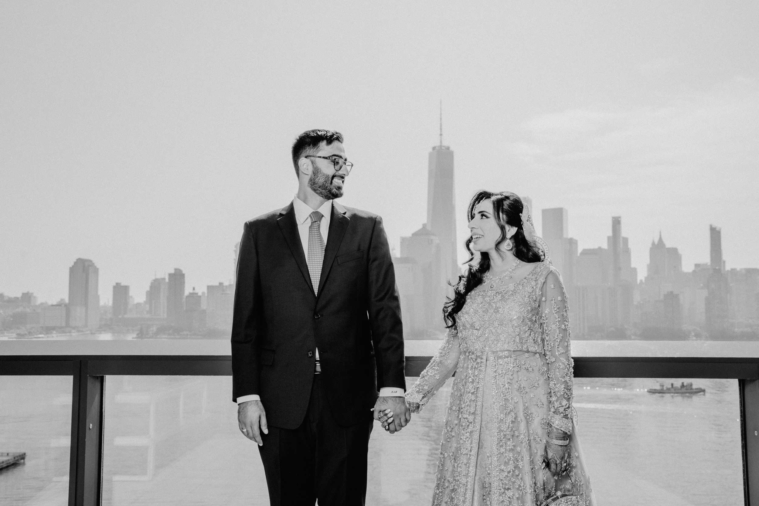 A couple dressed in wedding attire holding hands, standing on a balcony with the New York City skyline in the background, including the One World Trade Center, in black and white.