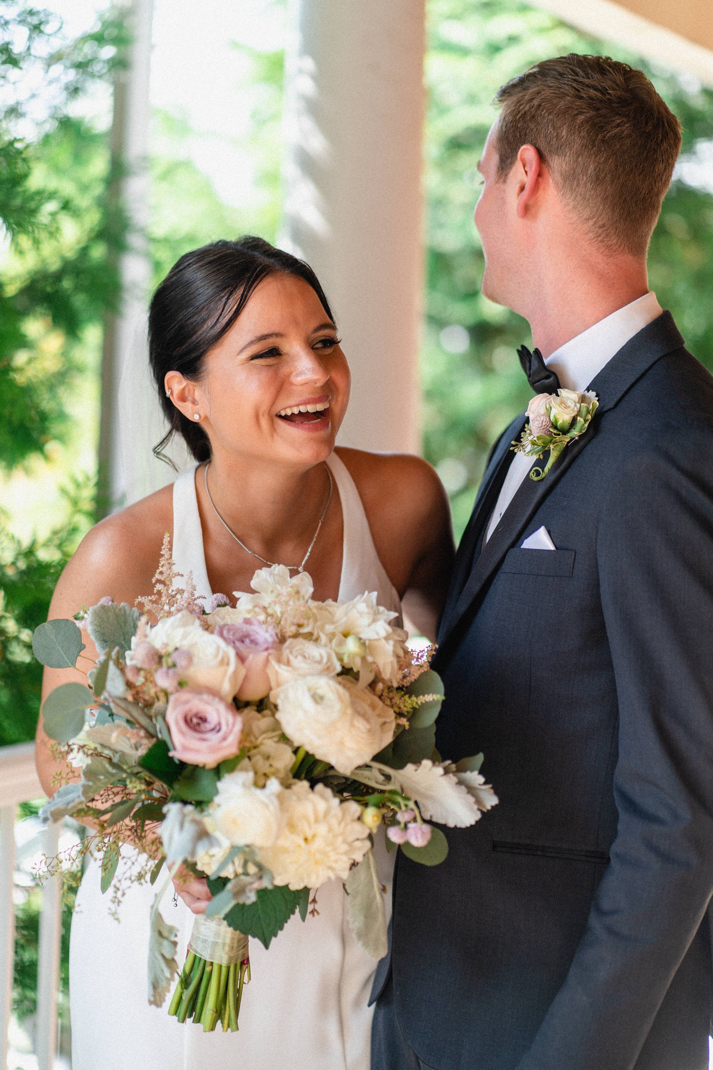 A bride and groom sharing a joyful moment on their wedding day, with the bride holding a large bouquet of flowers and the groom wearing a tuxedo with a boutonniere.