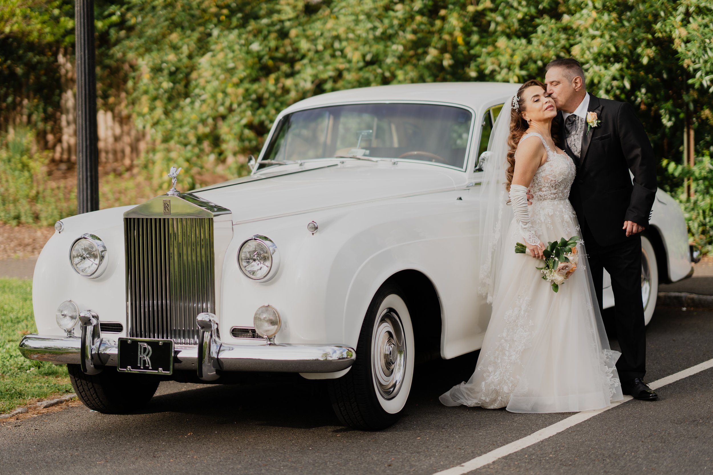 Bride and groom in wedding attire sharing a kiss next to a vintage white Rolls Royce car.