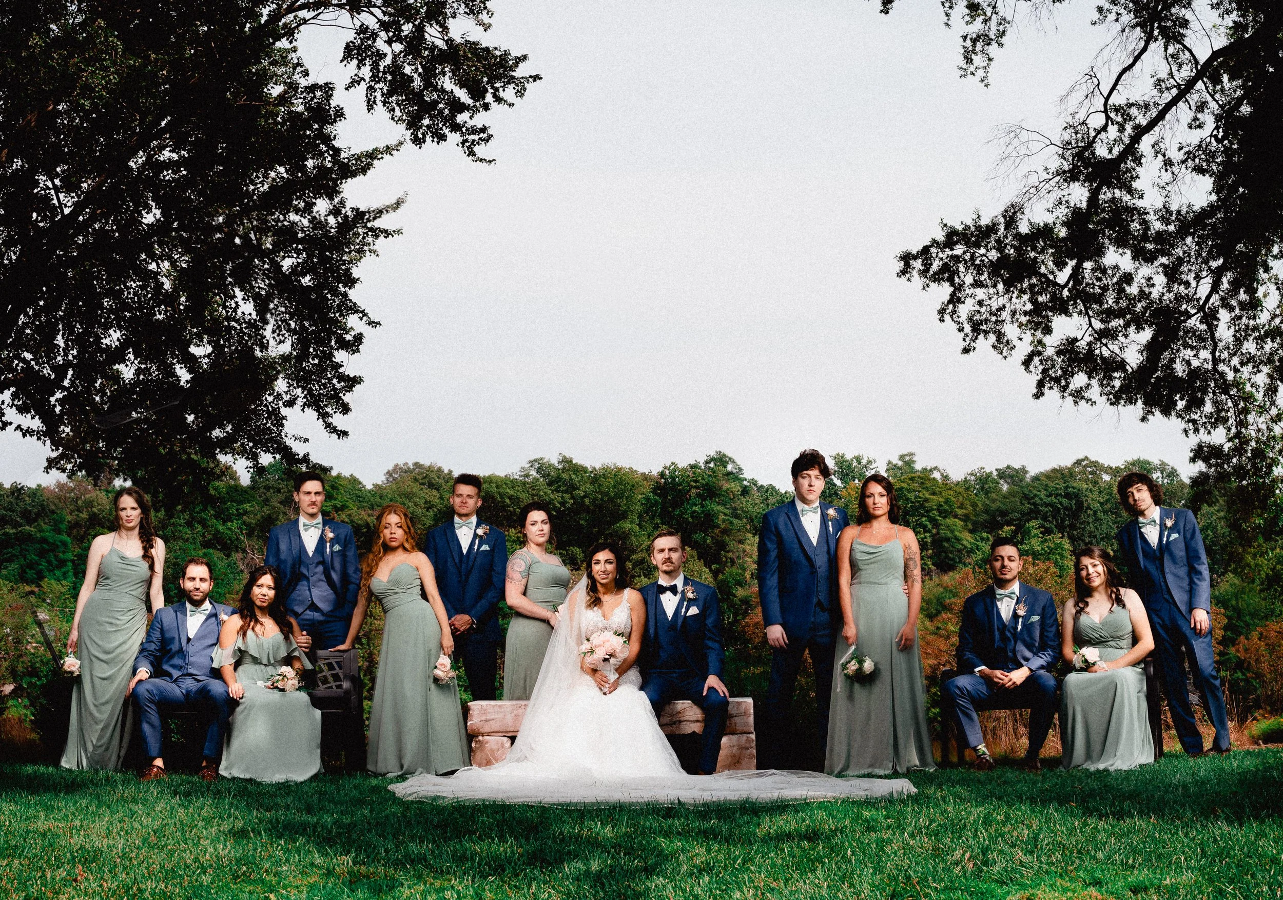 Editorial wedding photography portrait of a formal wedding party standing outdoors in a tree-lined park beneath a cloudy sky.