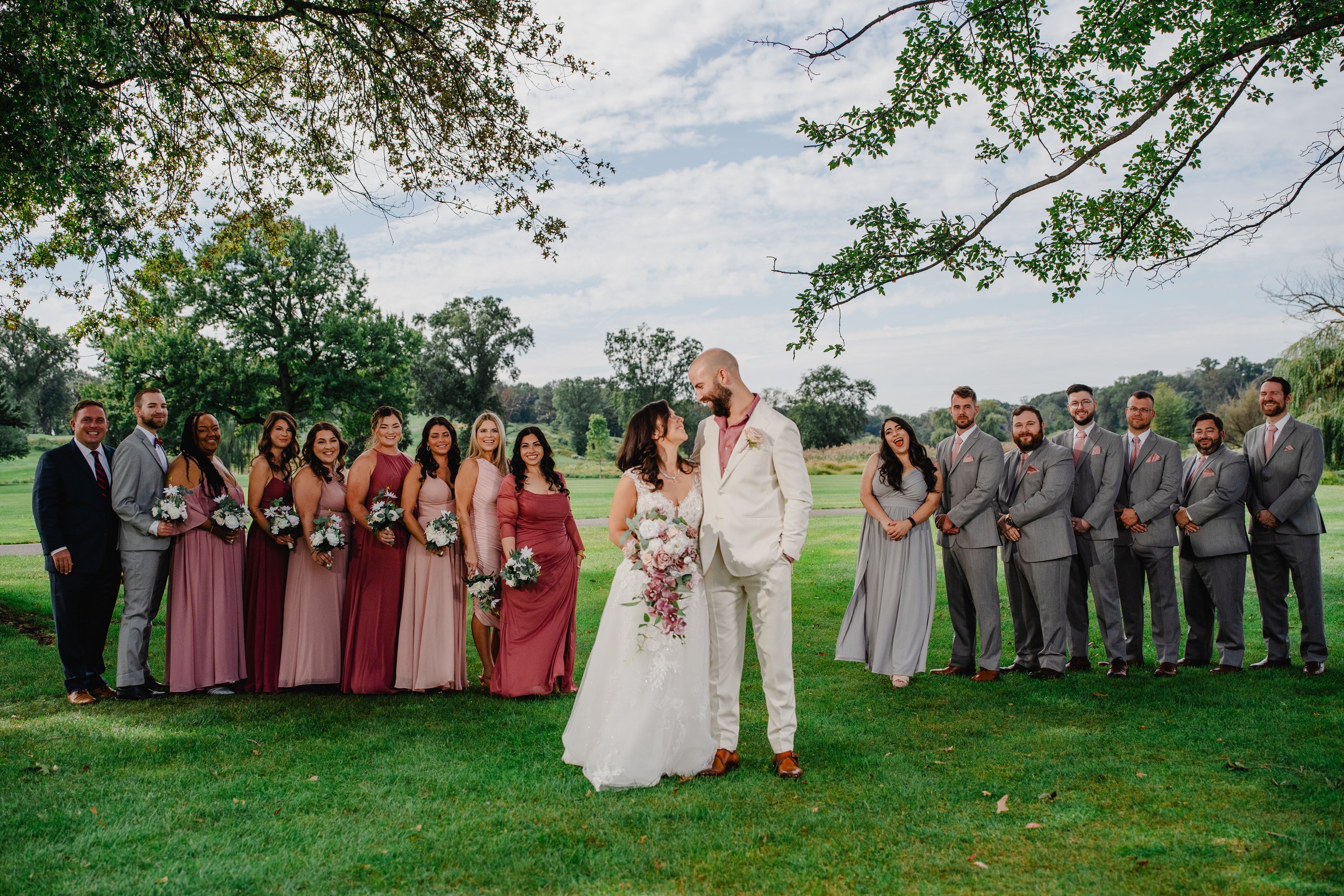 Group of wedding party members, including bride and groom, standing on a grassy field outdoors with trees and cloudy sky in the background.