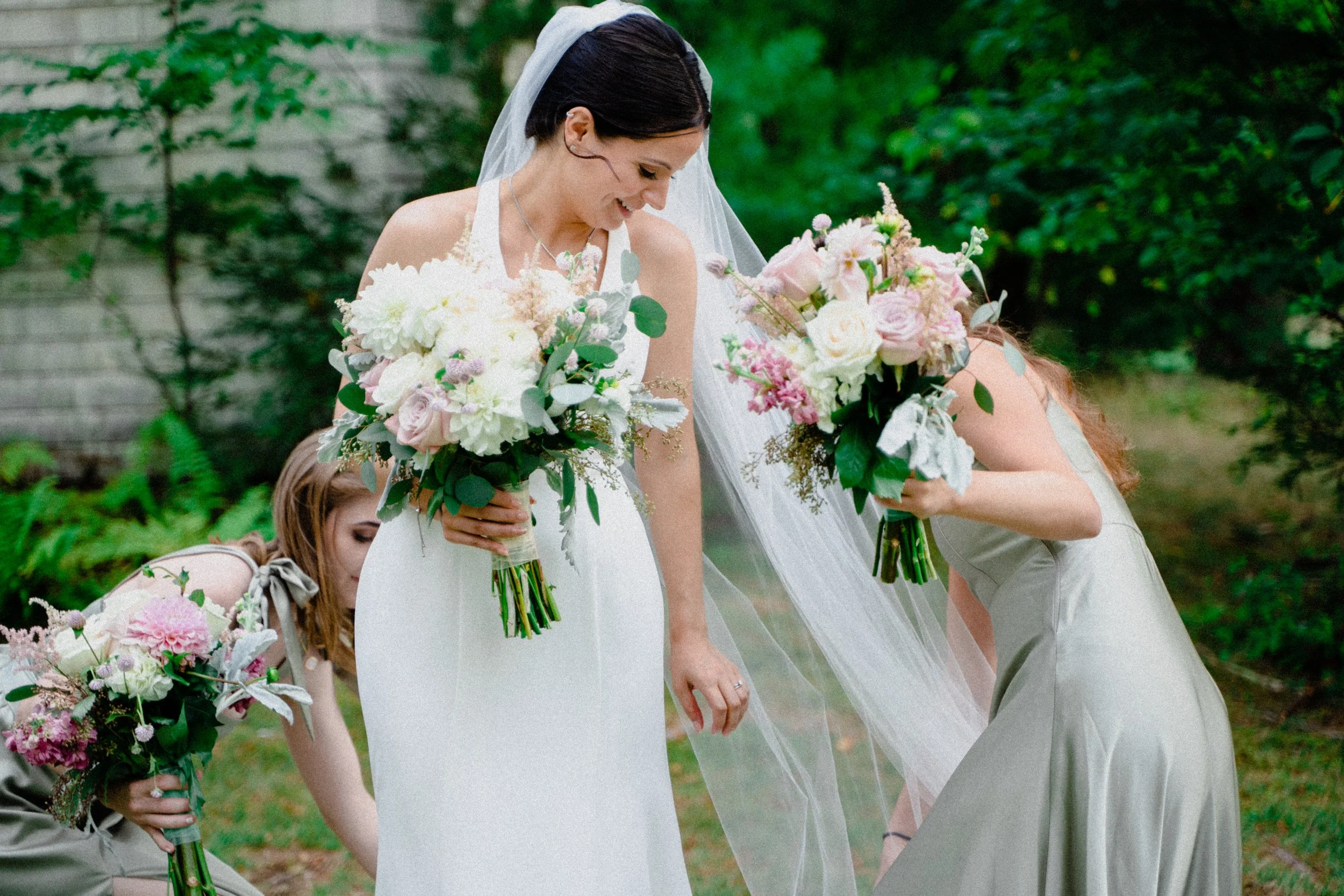 A bride in a white dress and veil holding a bouquet of white and pink flowers, smiling while standing outdoors, surrounded by women in gray dresses holding similar bouquets.