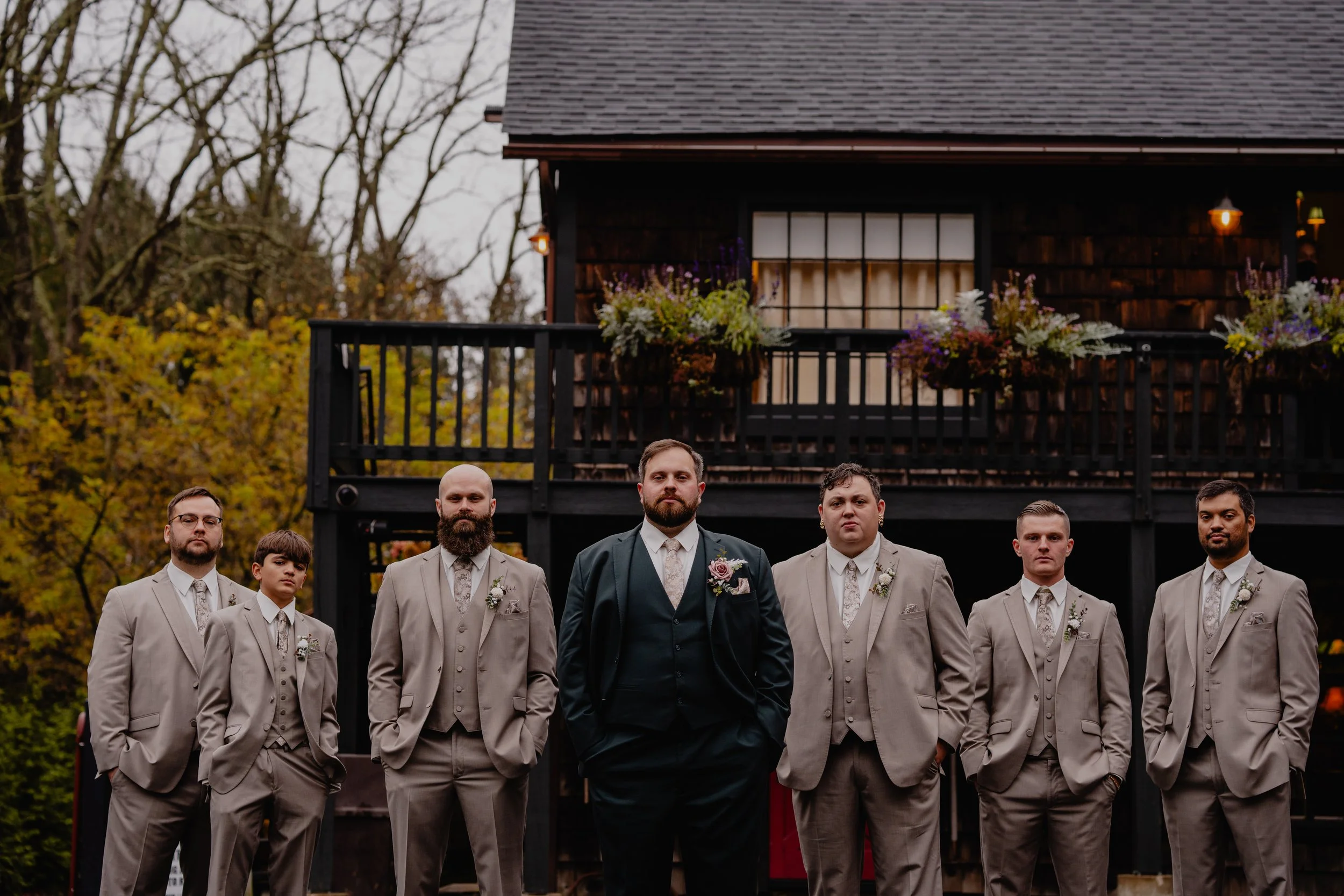 Groom in a black suit with a pink boutonniere and five groomsmen in beige suits with floral boutonnieres, standing in front of a dark wooden house with flower arrangements on the balcony, amidst autumn trees.