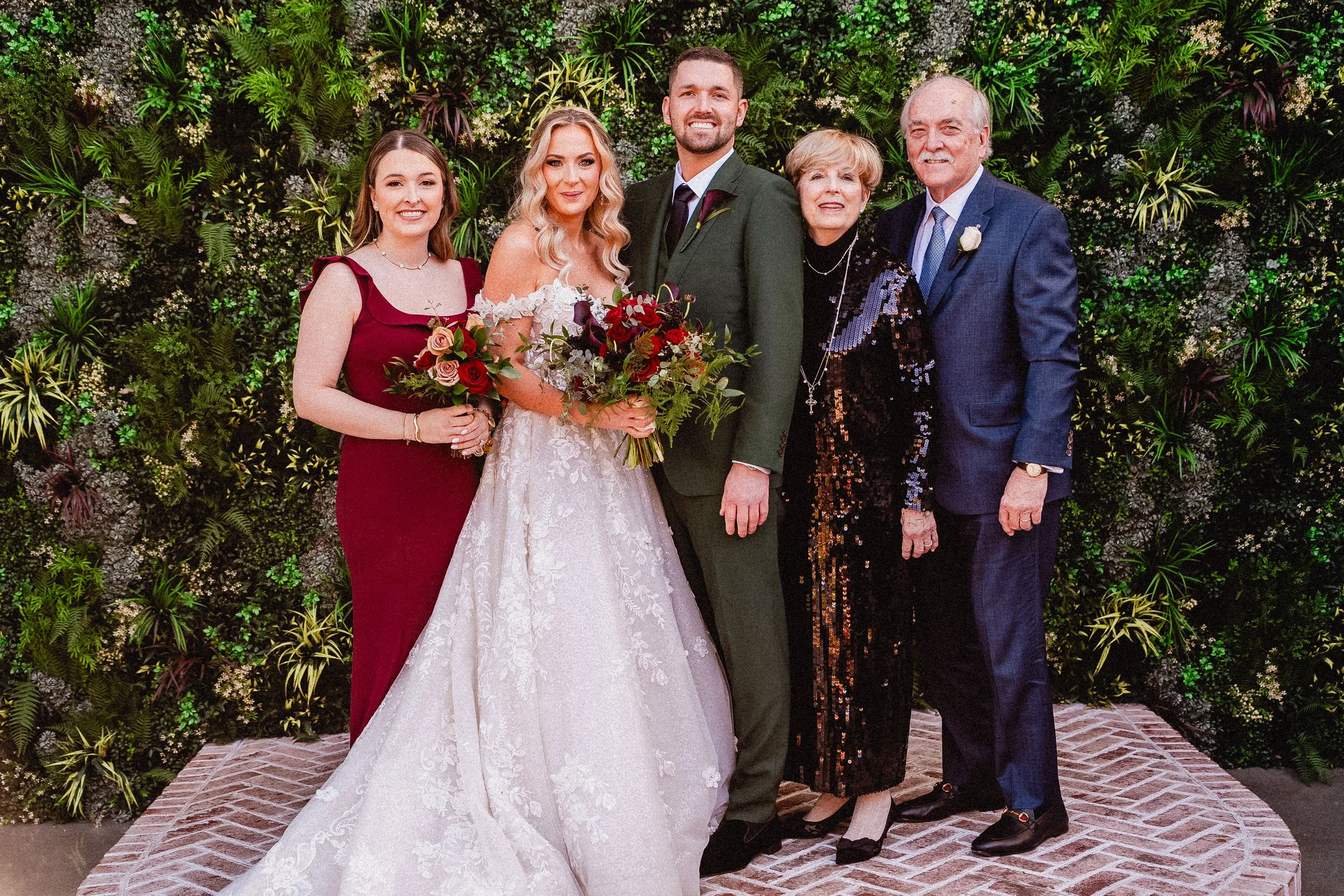 A group of six people in formal attire standing on a patterned brick floor in front of a lush green floral wall, celebrating a wedding.