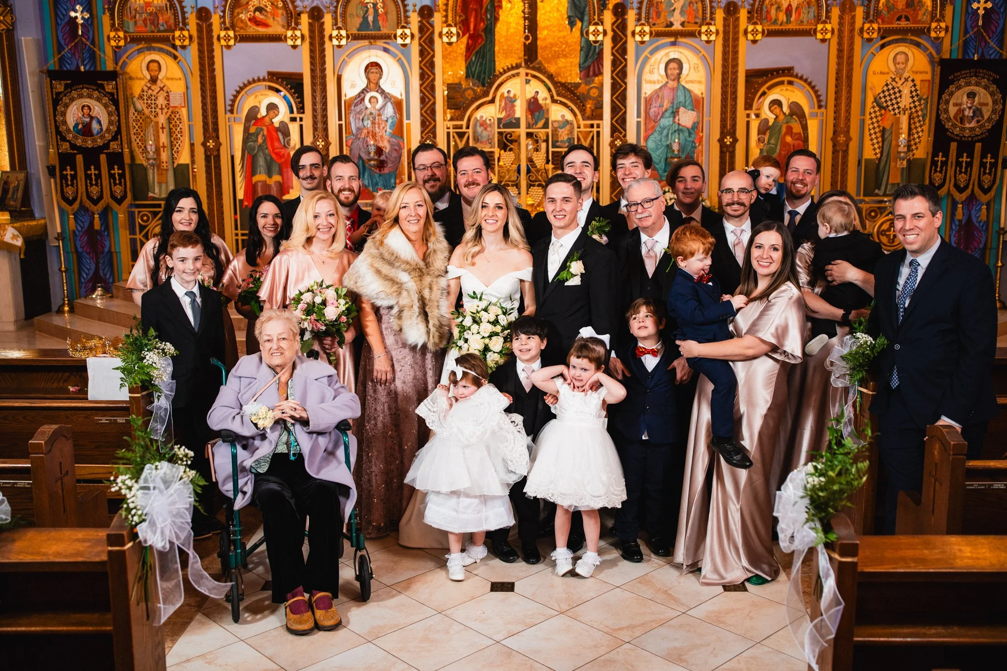 A group of people, including a bride and groom, family, and children, gathered for a wedding photo inside a church with colorful religious icons on the wall.
