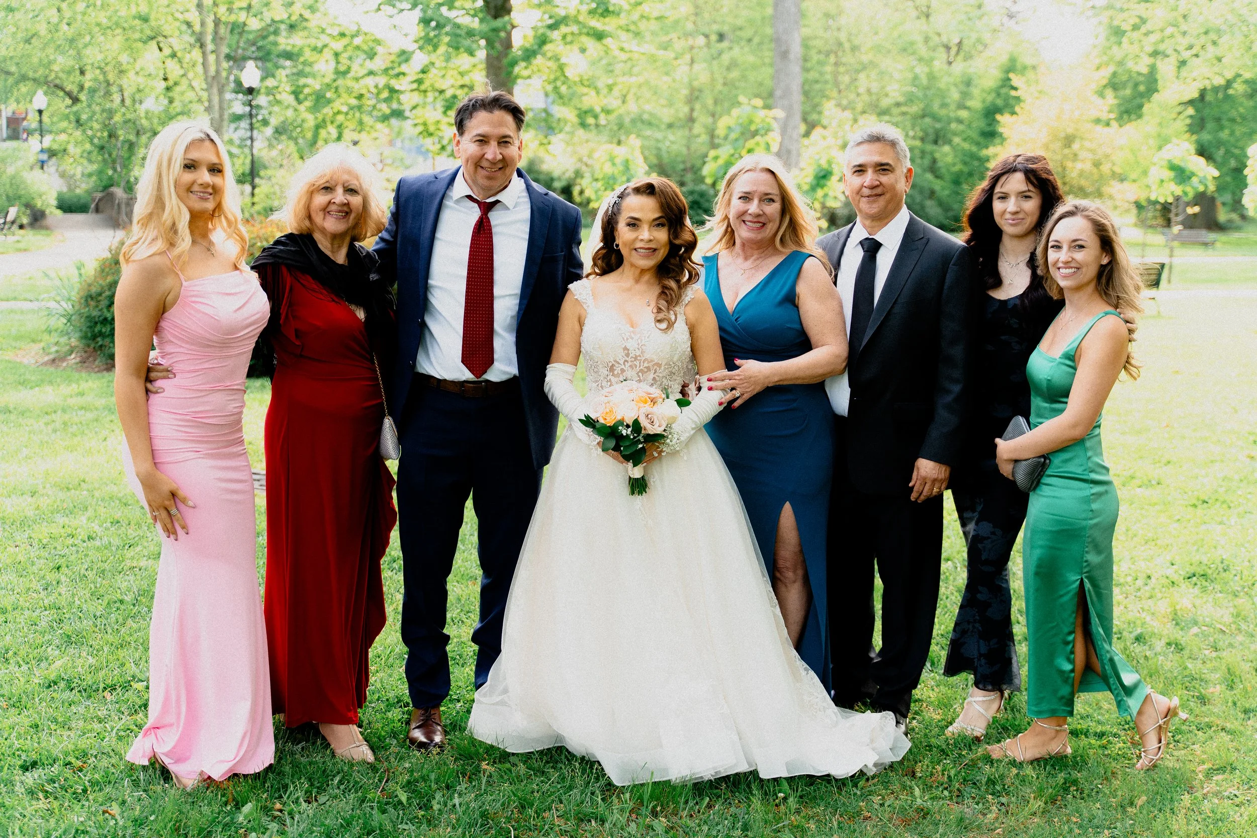 Group of eight people, including a bride in a white gown holding a bouquet, standing outdoors in a park with trees, smiling for a wedding photo.