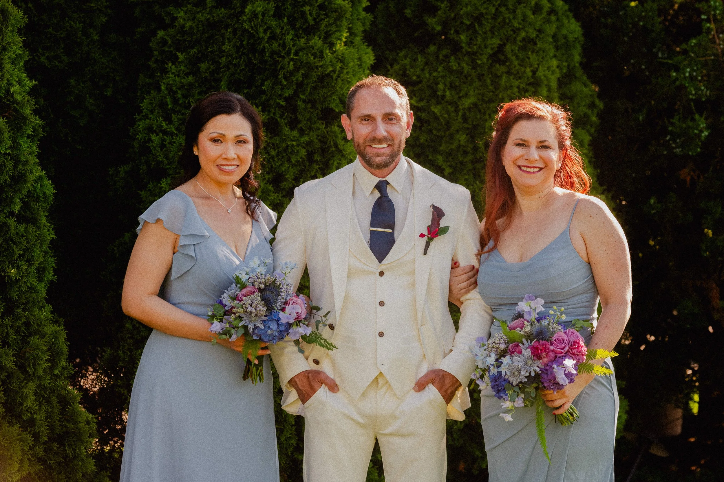 A man in a cream suit with a dark tie and a boutonniere stands between two women in matching light blue dresses, all smiling and holding bouquets of purple, pink, and white flowers, outdoors with greenery in the background.