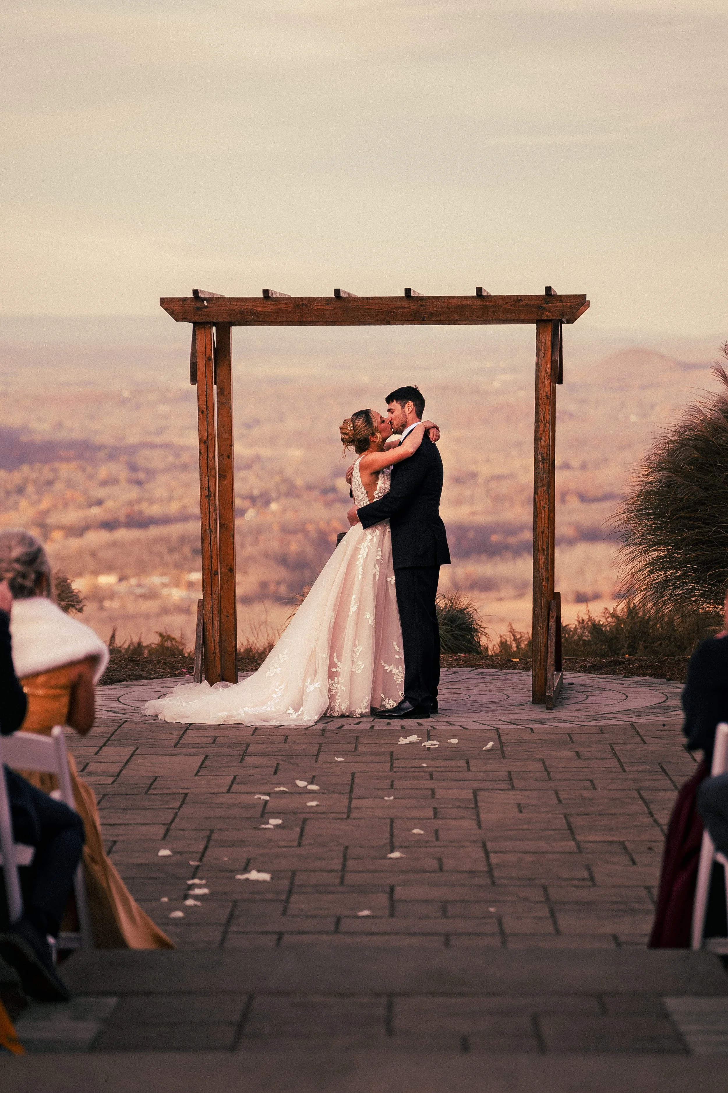 A bride and groom share a kiss during their outdoor wedding ceremony at sunset, surrounded by seated guests and a scenic landscape in the background.