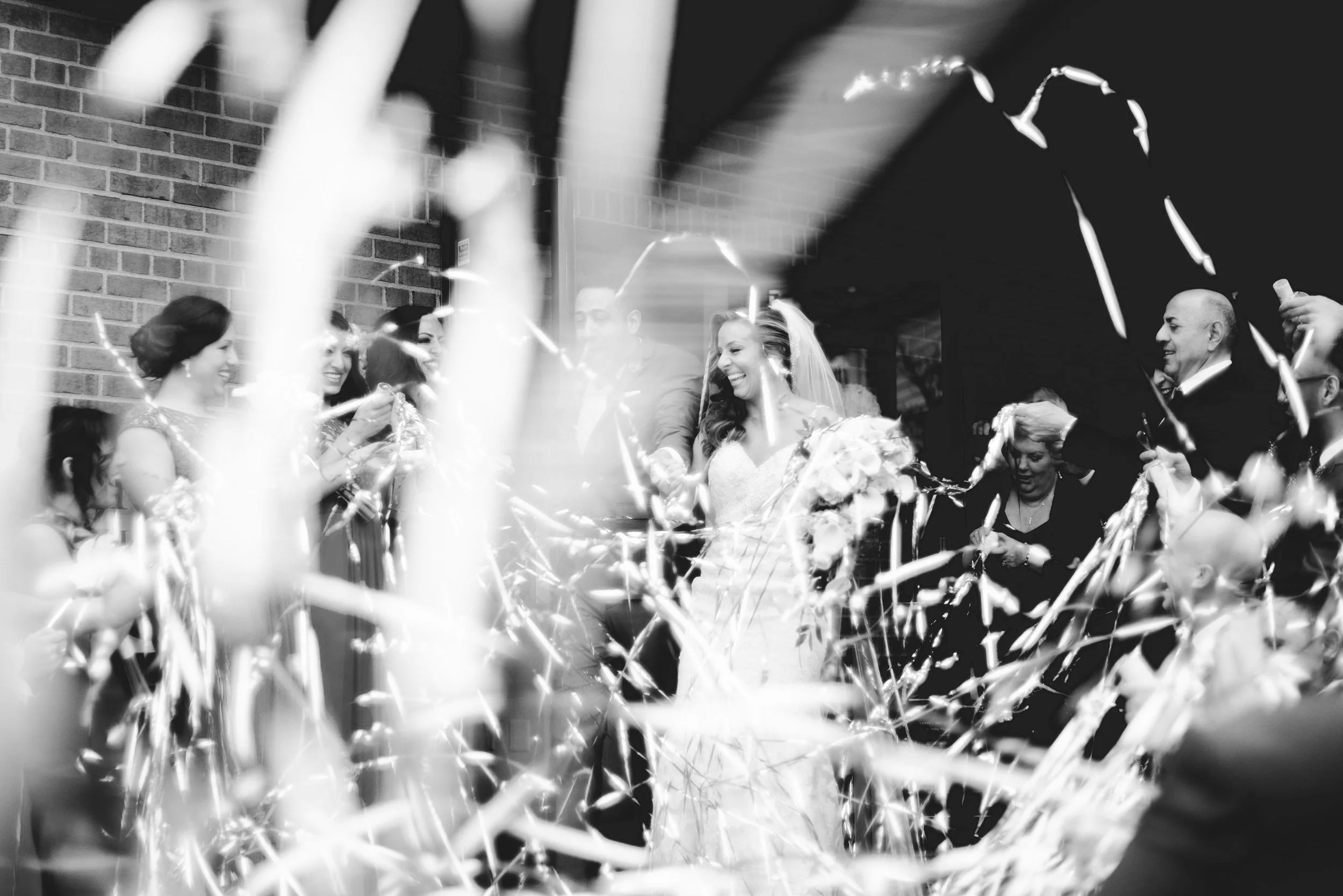 Black and white photo of a wedding celebration with a bride holding flowers, surrounded by companions, with confetti falling around them.