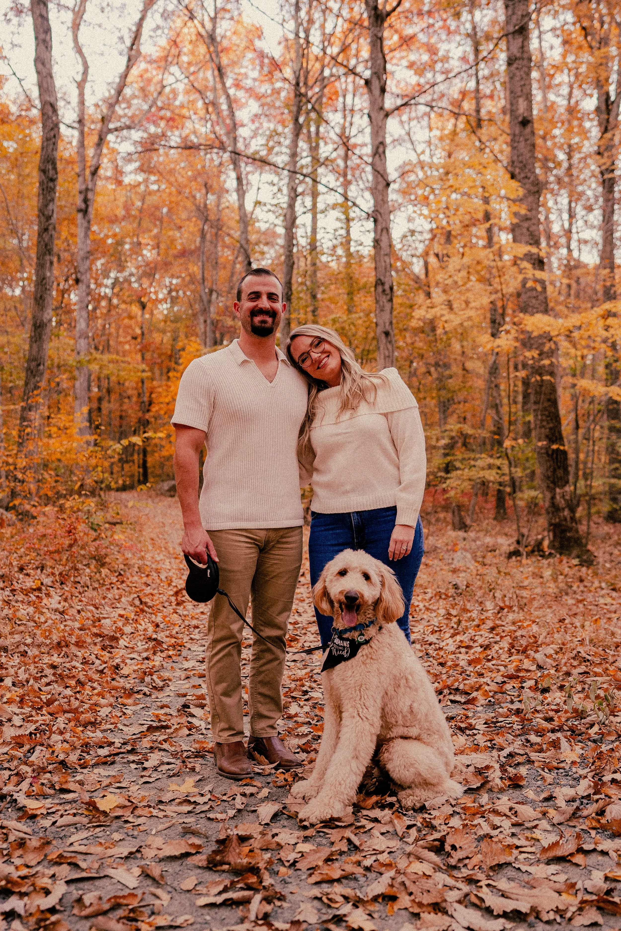 NJ Wedding Photography of A smiling couple with a golden doodle dog in an autumn forest with orange leaves.