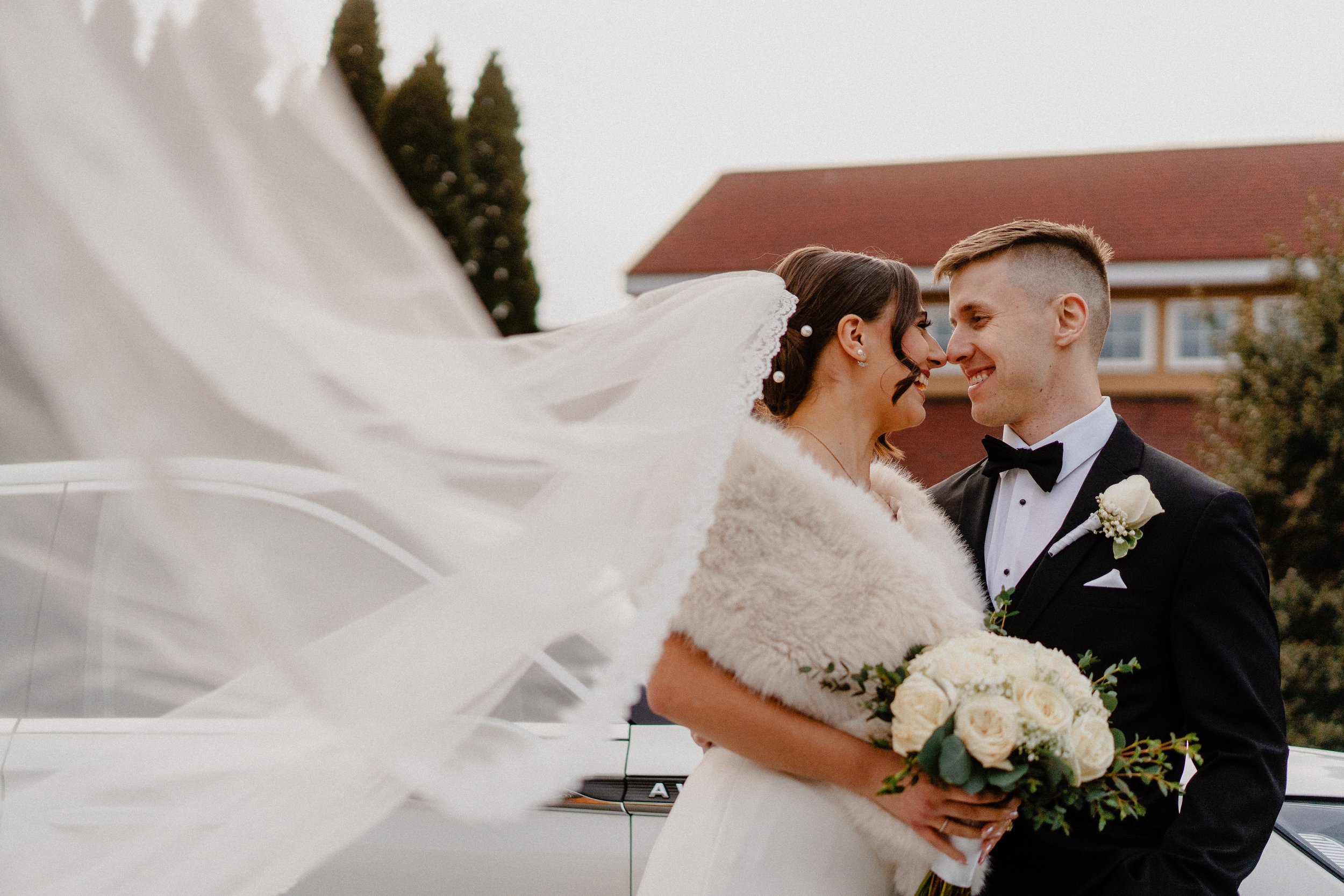 A newlywed couple smiling and touching foreheads outdoors, the bride holding a bouquet of white roses, the groom dressed in a black tuxedo with a white rose boutonniere, a white wedding veil is flowing in the breeze.