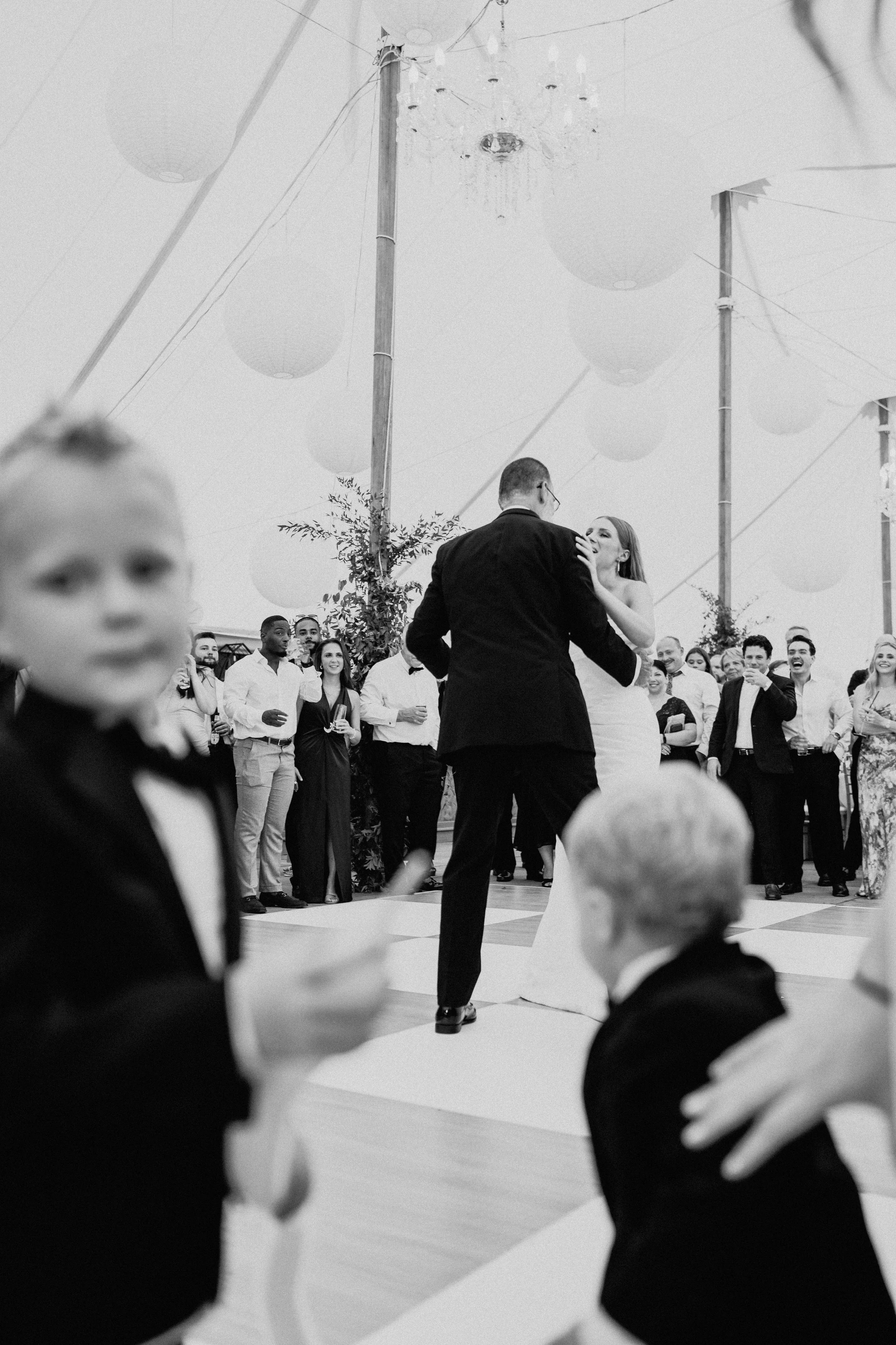 Black-and-white documentary wedding photography of a child watching the groom dance beneath the reception tent.