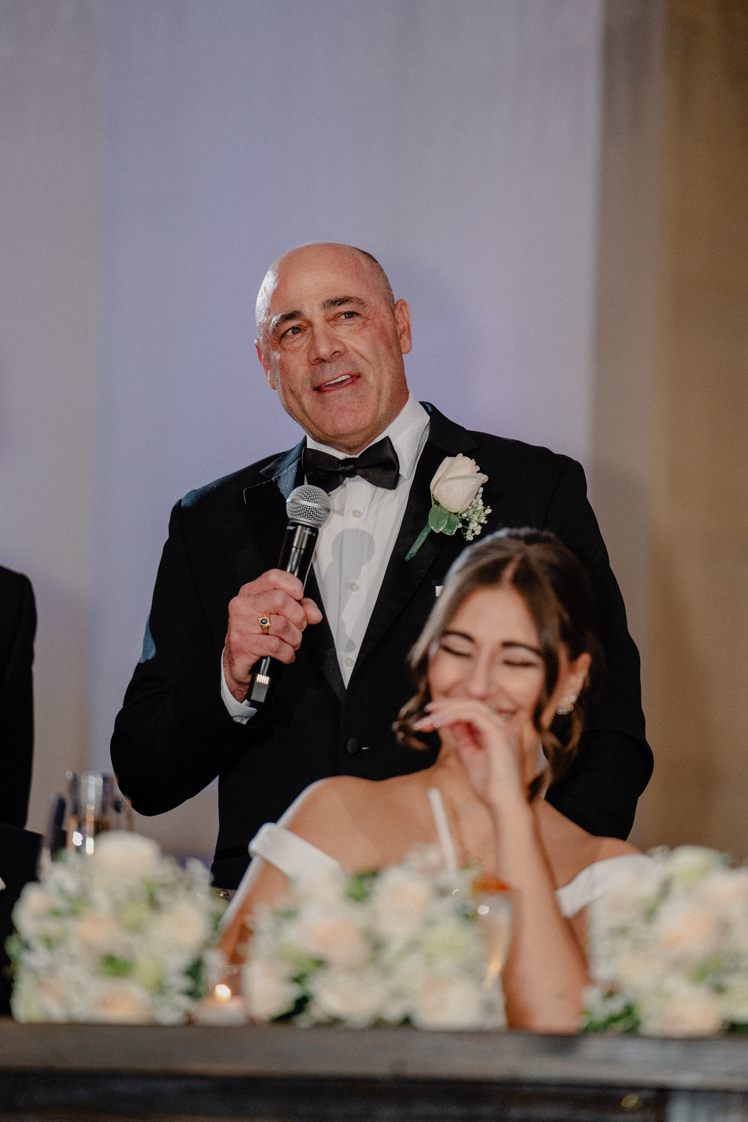 A man in a tuxedo holding a microphone, standing behind a smiling woman at a wedding reception table with floral arrangements.