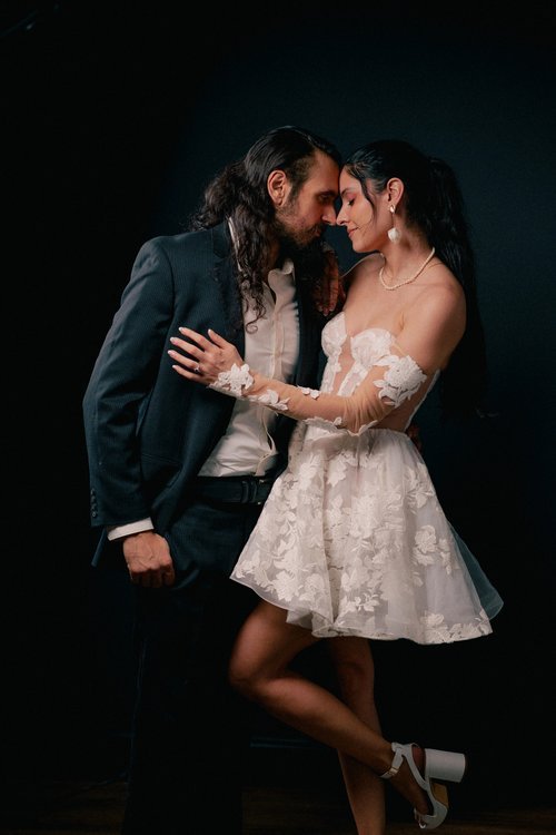 A man and woman standing close together with foreheads touching, dressed in formal attire, against a dark background.