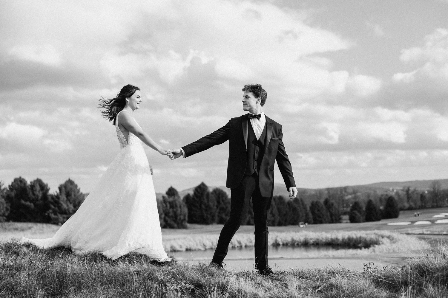 A bride and groom holding hands outdoors on a grassy hill, with trees and a cloudy sky in the background, in black and white.