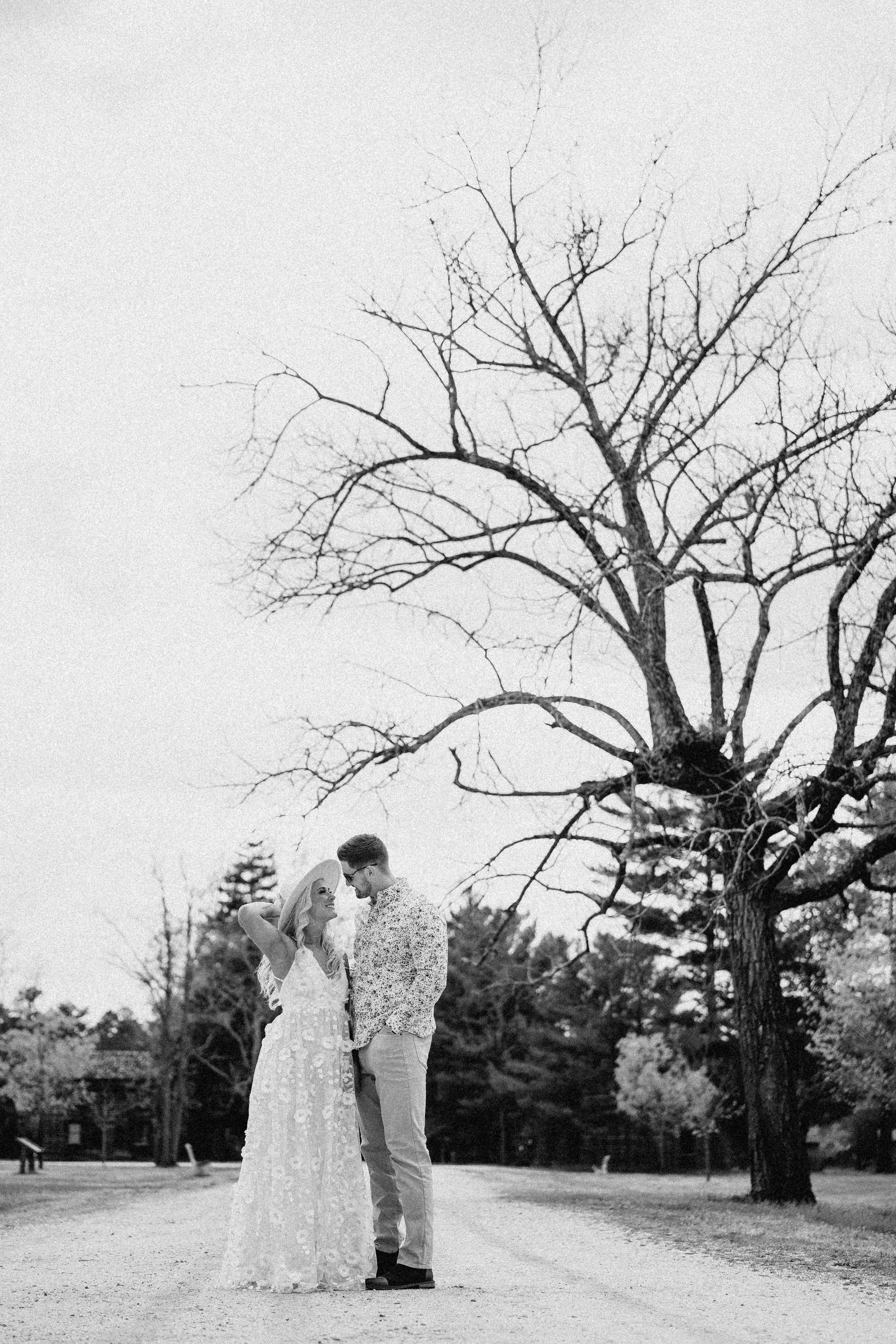 NJ Wedding Photography black and white photo of a couple standing close under a large, leafless tree in a park or open area, with the woman wearing a long, flowy dress and a wide-brimmed hat, and the man in a patterned shirt and light-colored pants.
