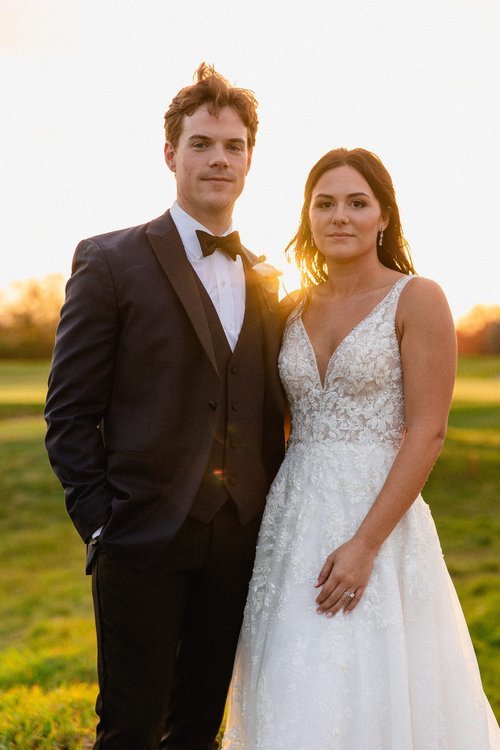 A bride and groom standing outdoors at sunset, the groom in a black tuxedo with a bow tie, and the bride in a white lace wedding dress.