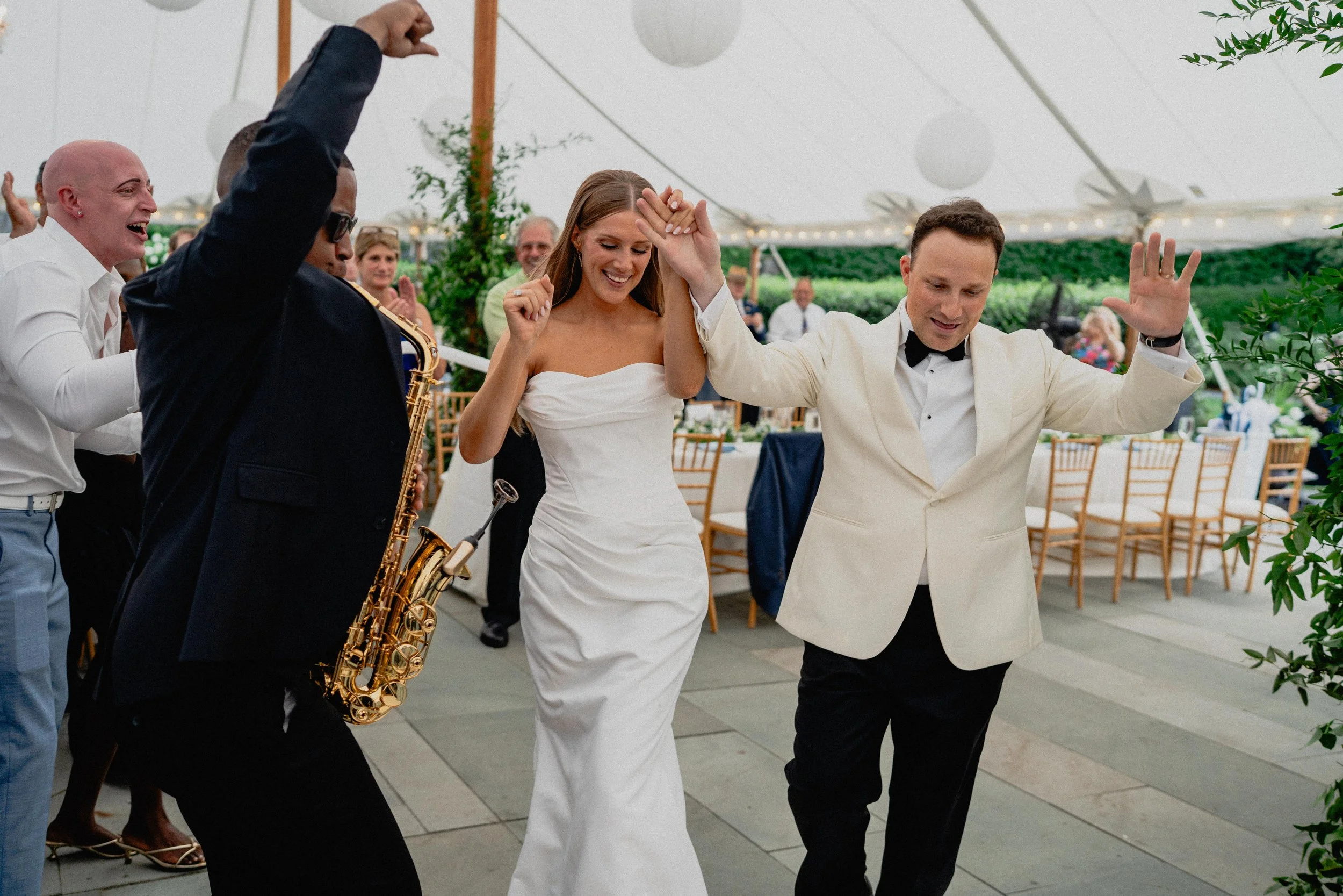 Documentary wedding photography of newlyweds entering the tent reception with raised hands as a saxophonist performs.