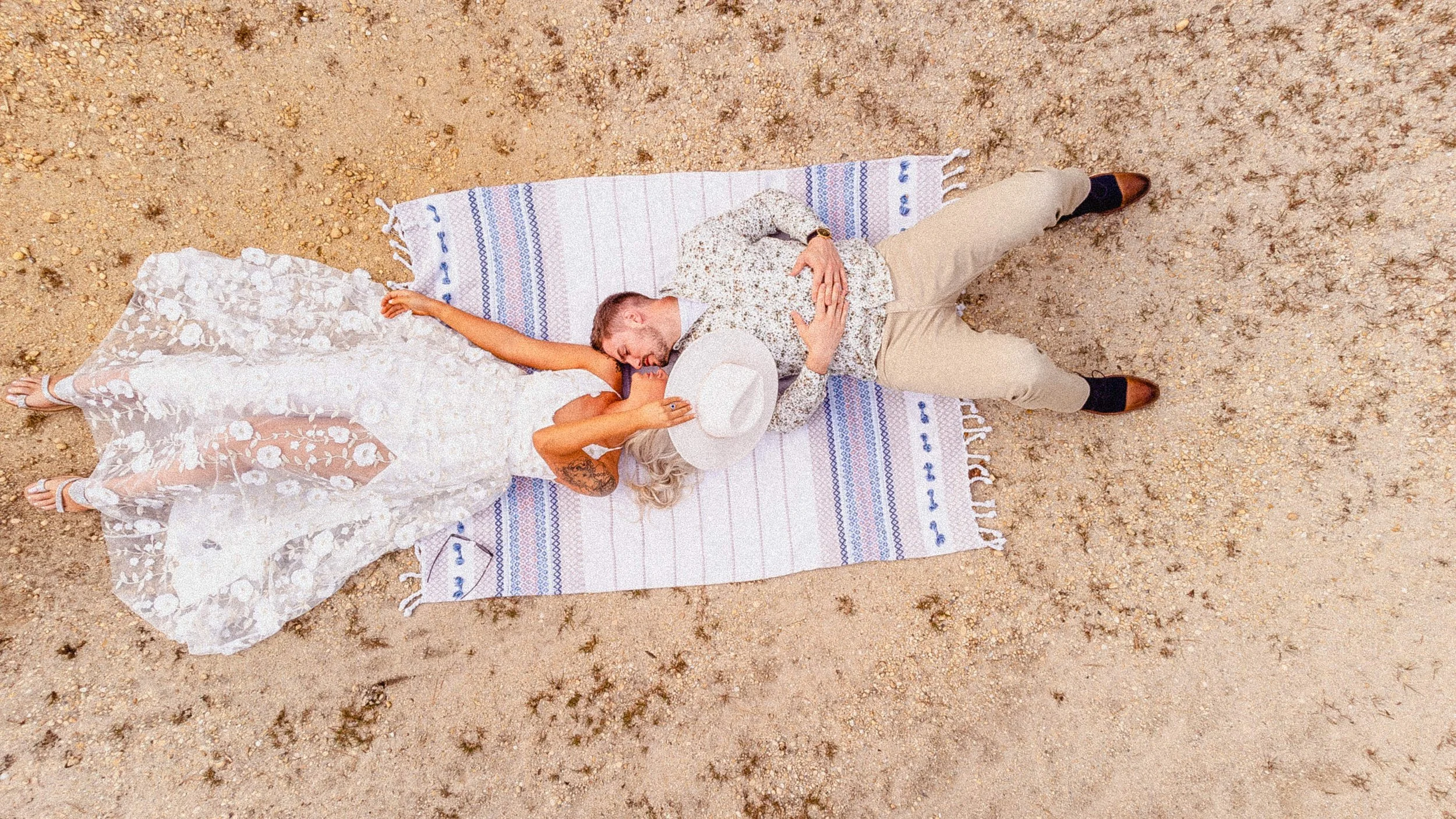 NJ Wedding Photography of A couple lying on a striped blanket on the beach at sunset, with the woman in a lace wedding dress and the man in a floral shirt and beige pants, sharing a tender moment.