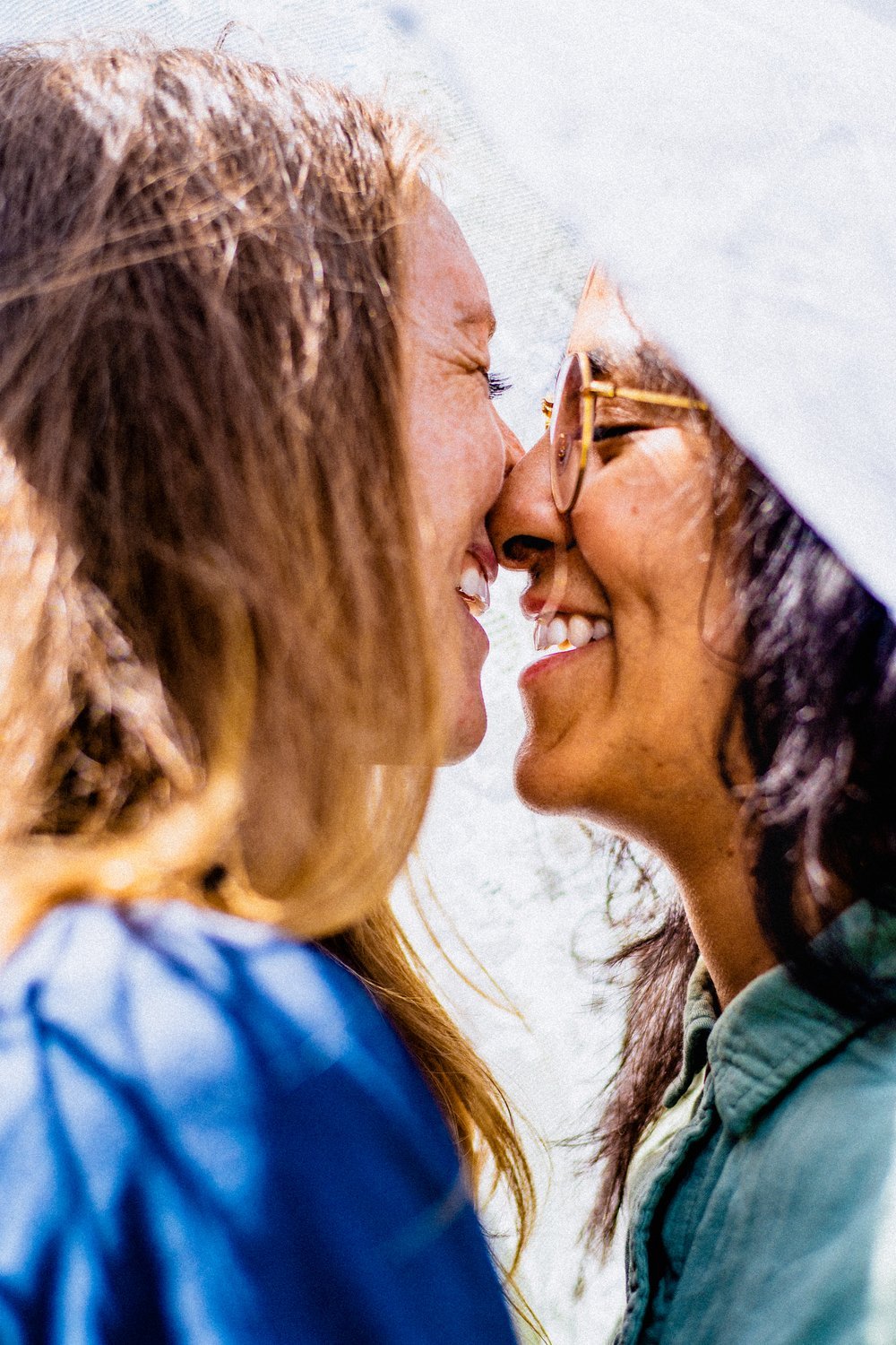 Two women are smiling and touching noses under a white umbrella, close together, enjoying a happy moment outdoors.