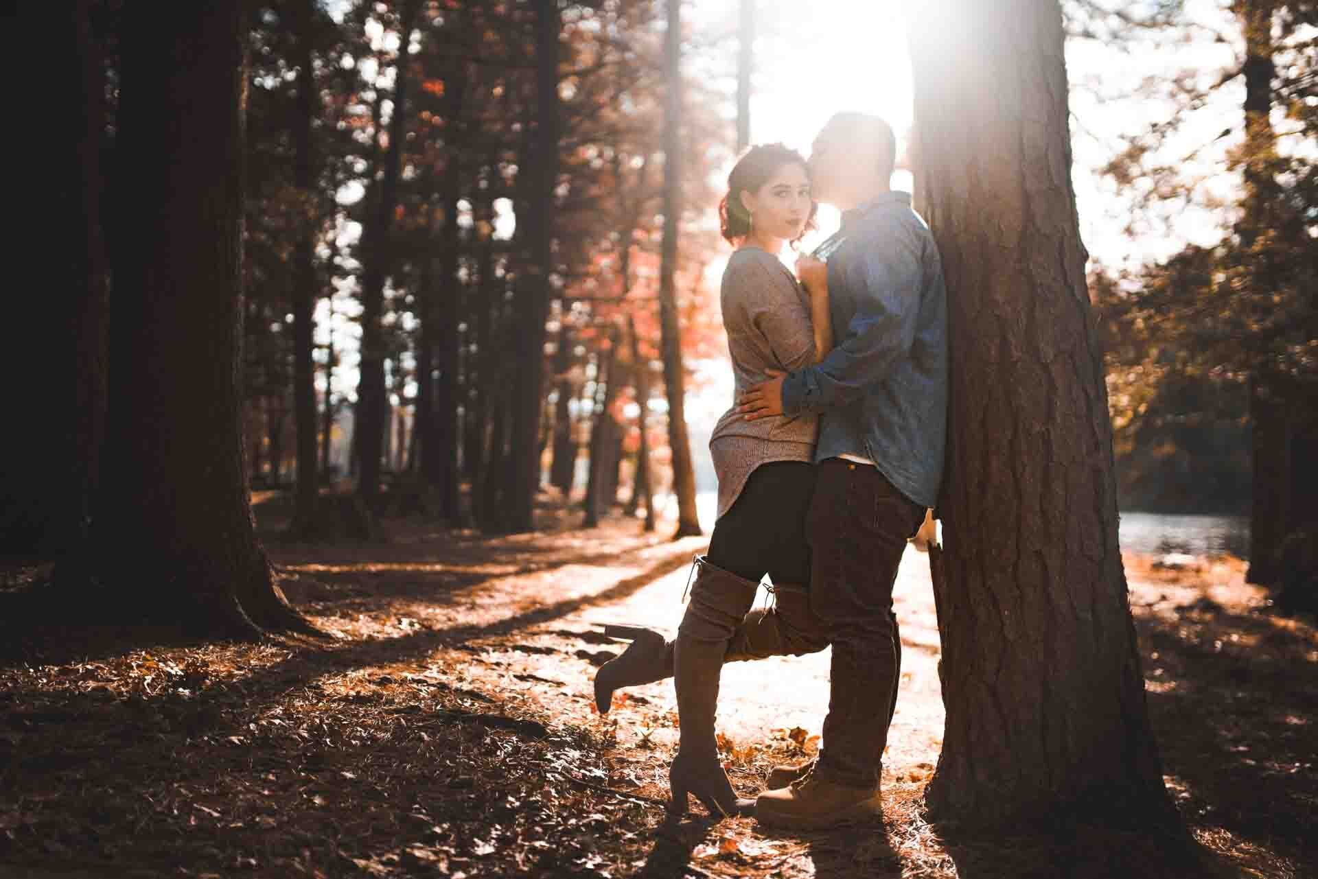 A couple standing close to each other behind a tree in a forest during sunset