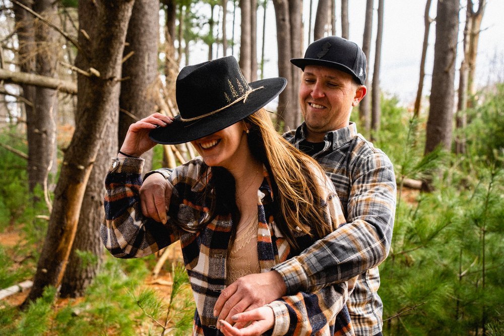 A smiling couple in plaid shirts enjoying a walk in a forest, with the woman wearing a black hat and the man holding her from behind.