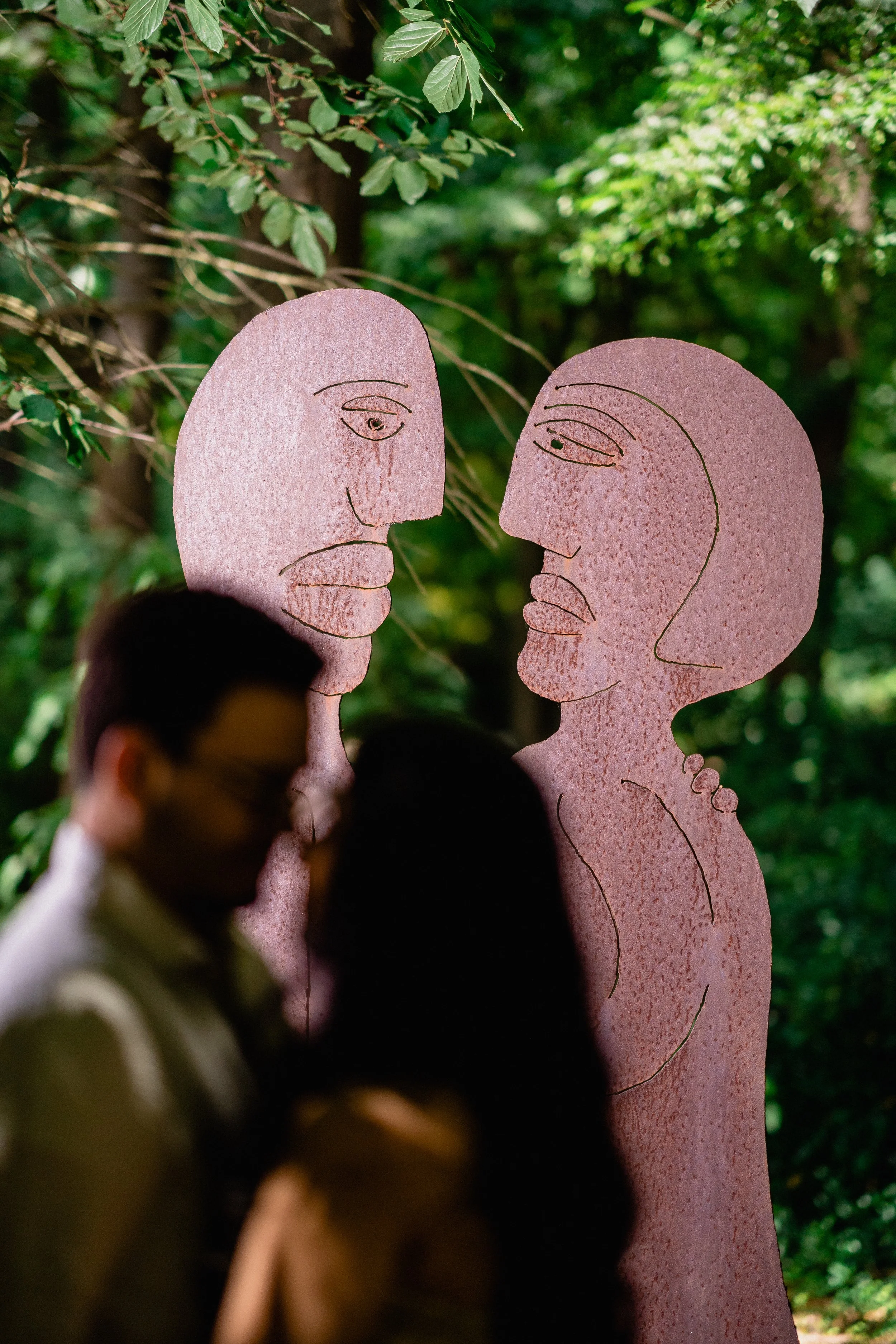 NJ Wedding Photography of Two pink abstract sculpture figures of a man and woman facing each other in a lush green outdoor setting, with two blurred people walking past in the foreground.