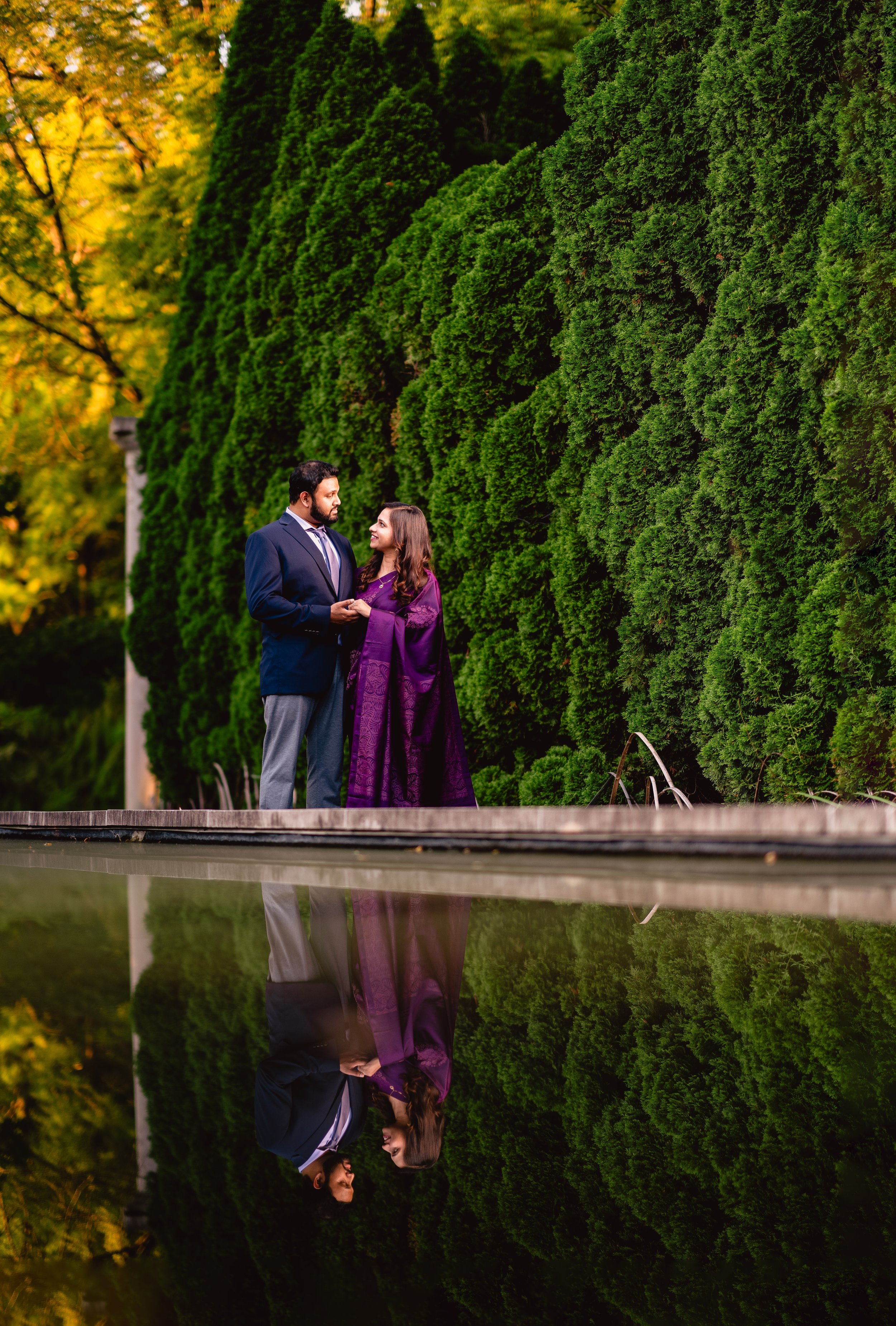 NJ Wedding Photography of A couple standing outdoors beside a tall green hedge during autumn, holding hands and looking at each other, with their reflection visible in a nearby water feature.